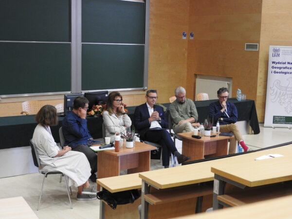 6 osób siedzi na krzesłach na scenie w auli i rozmawia, biorąc udział w panelu dyskusyjnym podczas konferencji.