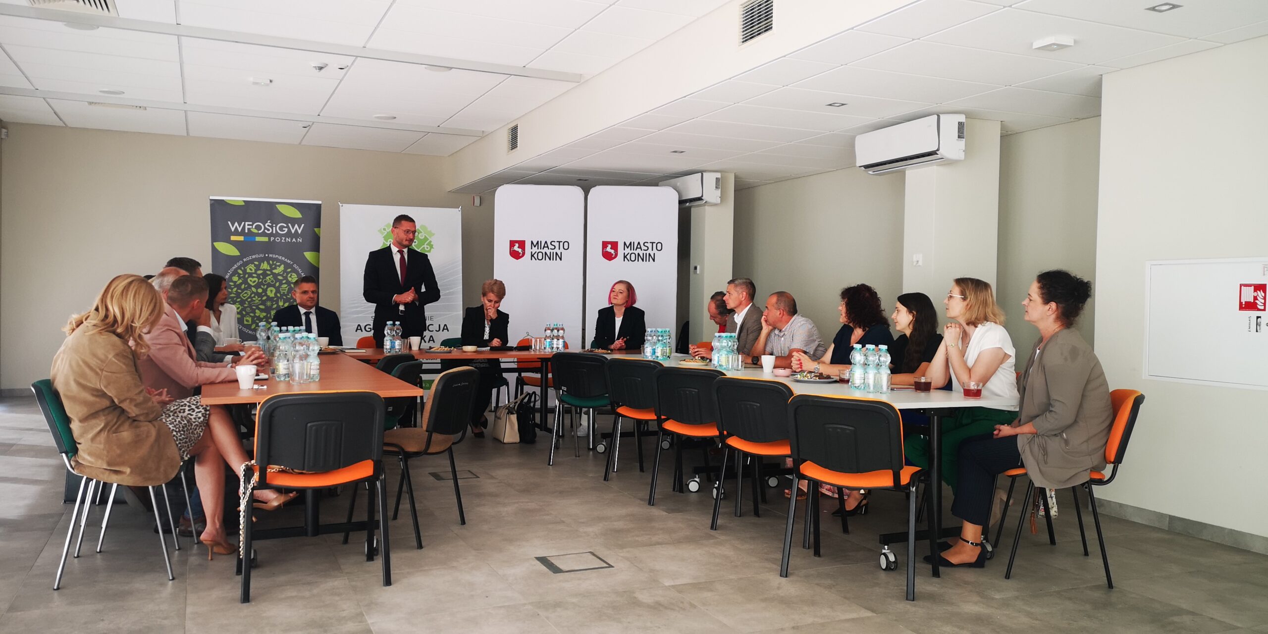 Grupa osób siedzi na spotkaniu za stołami na tle bannerów promocyjnych i omawia współpracę.