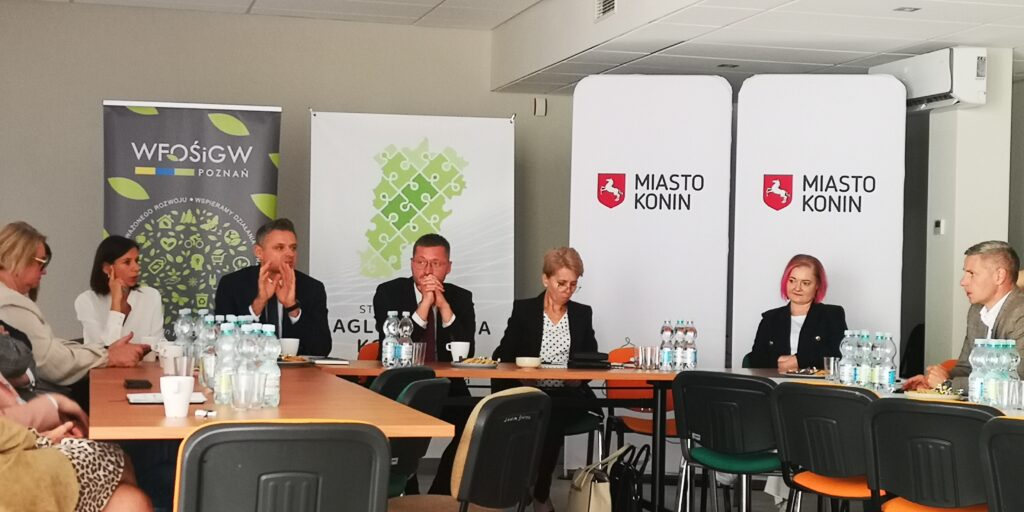 Grupa osób siedzi na spotkaniu za stołami na tle bannerów promocyjnych i omawia współpracę.