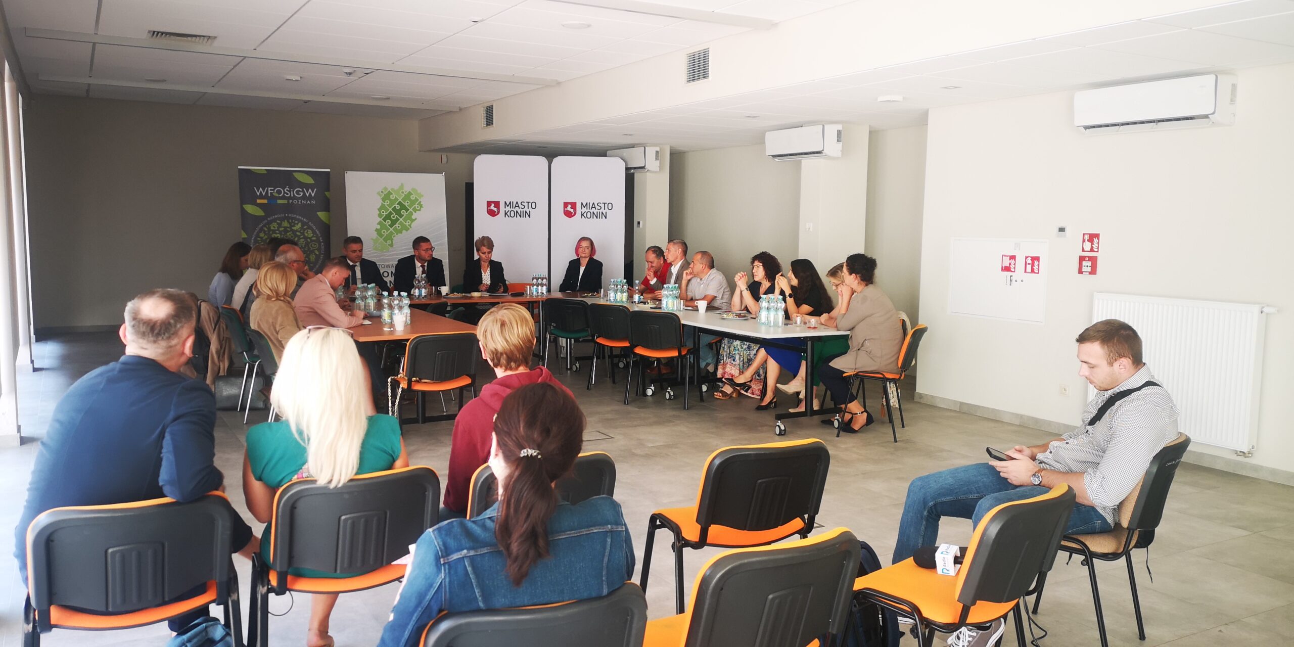 Grupa osób siedzi na spotkaniu za stołami na tle bannerów promocyjnych i omawia współpracę.