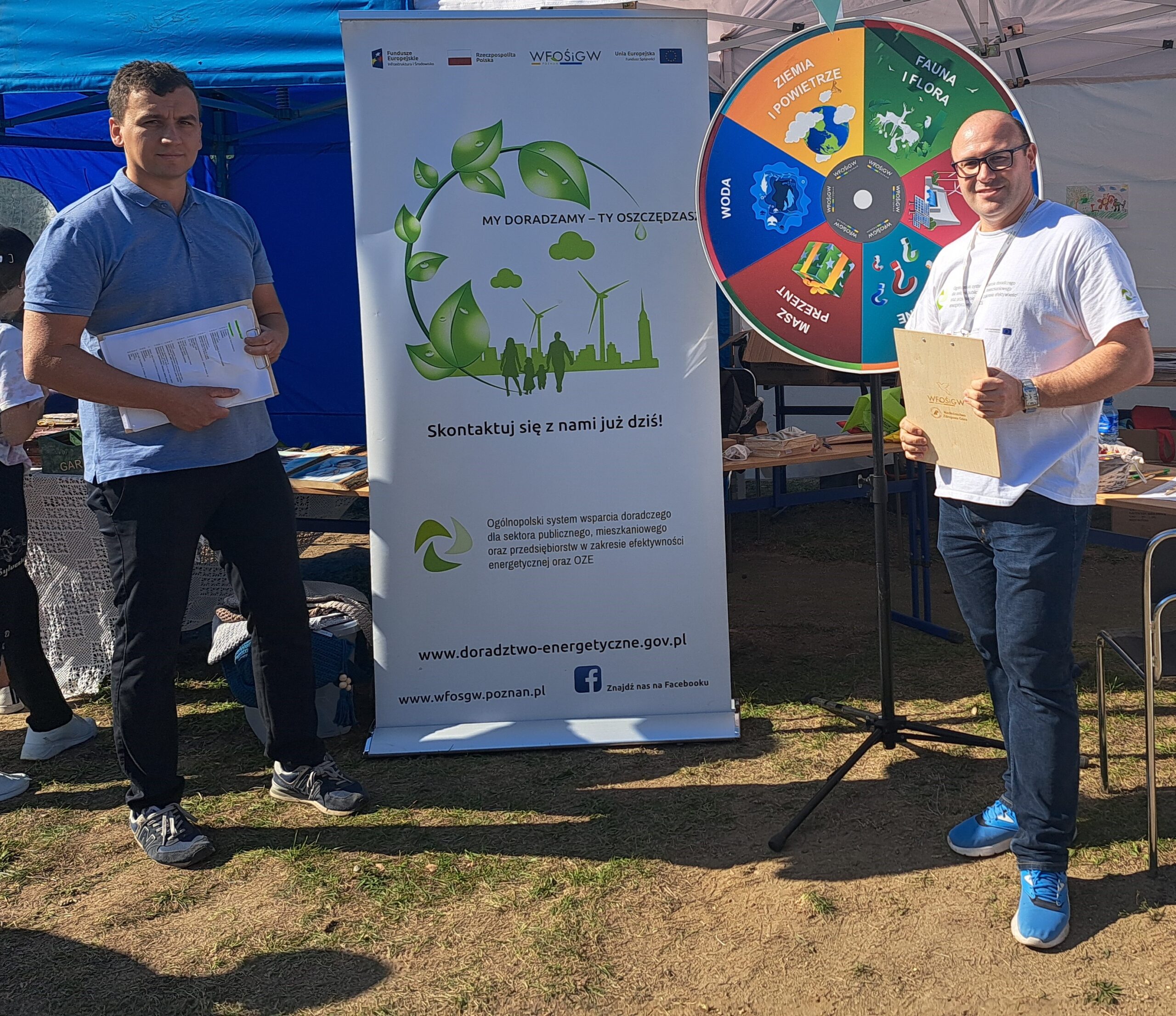 Dwóch doradców stoi przed stoiskiem podczas festynu, za nimi stoi koło fortuny.