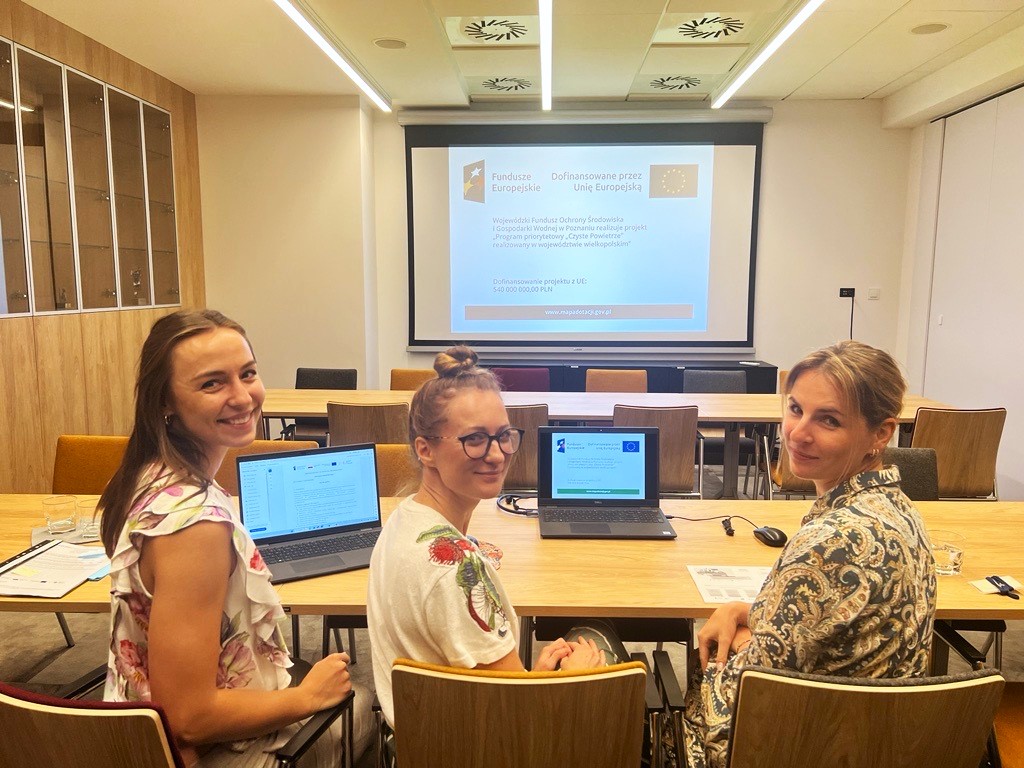 3 osoby siedzą przodem do obiektywu za stołem przed laptopem i prowadzą szkolenie online, w tle widać wyświetloną na dużym ekranie prezentację multimedialną.