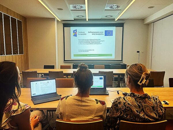 3 osoby siedzą tyłem do obiektywu za stołem przed laptopem i prowadzą szkolenie online, w tle widać wyświetloną na dużym ekranie prezentację multimedialną.