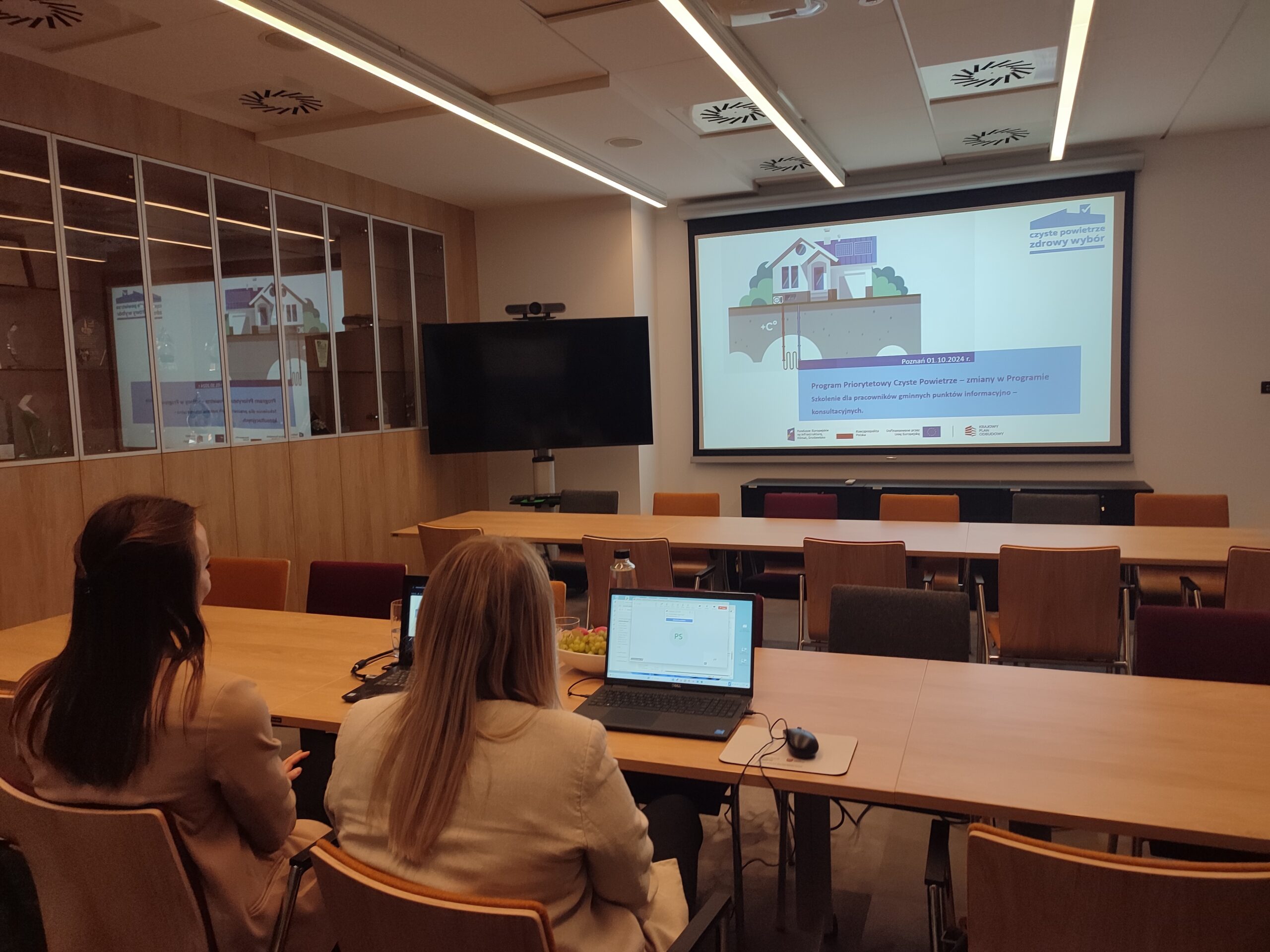 2 osoby siedzą przy stole naprzeciw wyświetlonej prezentacji i przeprowadzają szkolenie online.