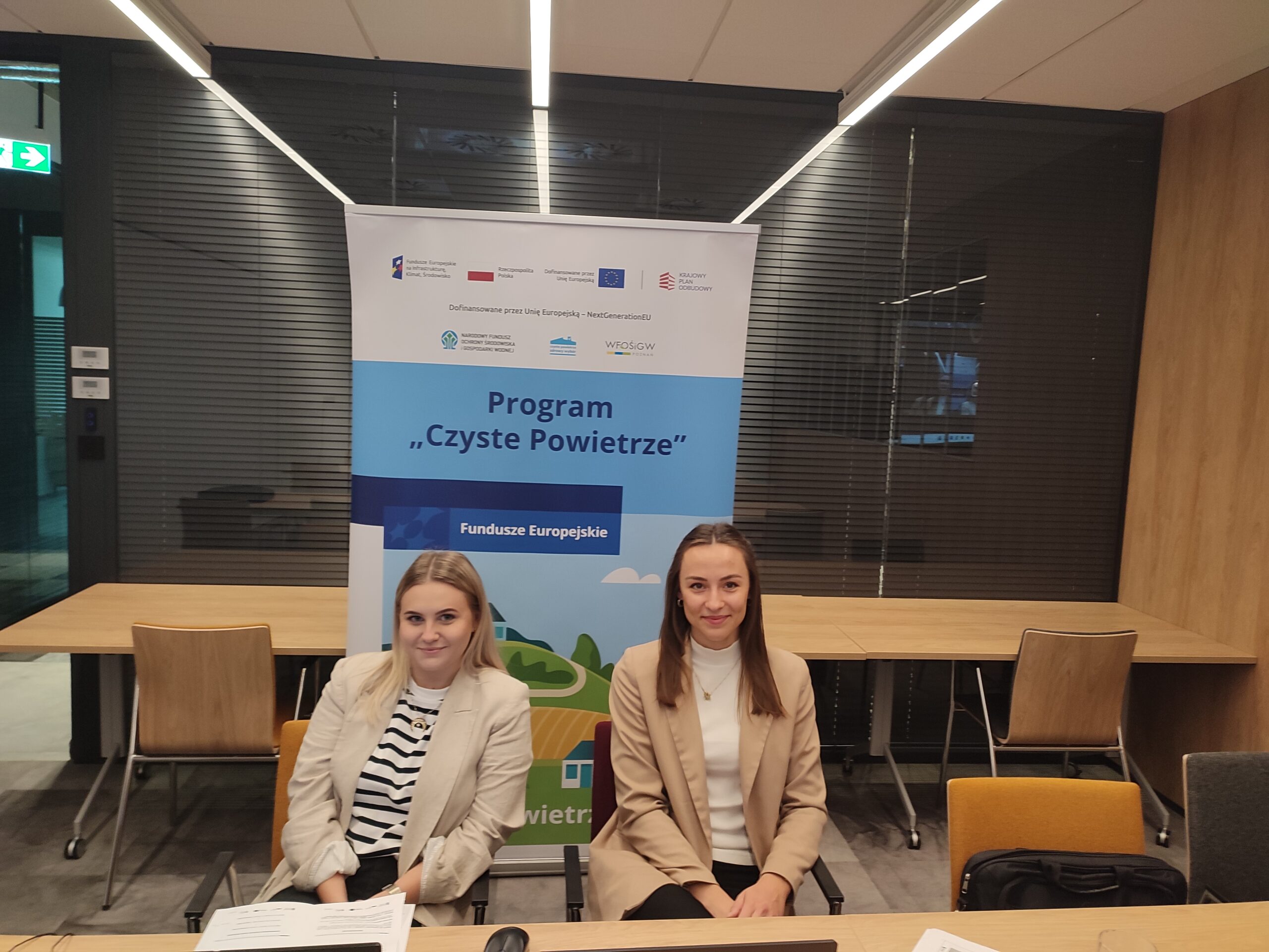 2 osoby siedzą za stołem, na którym stoją laptopy, a za nimi znajduje się rollup.