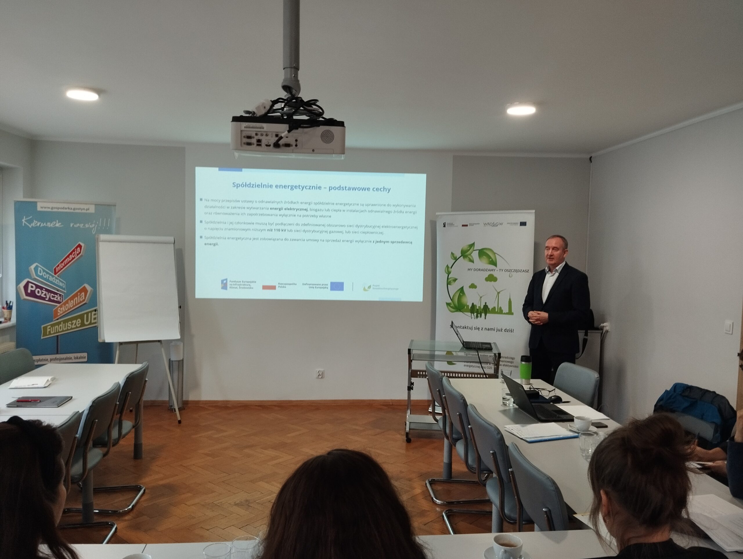 Na środku widać ekran projektora, na którym wyświetlana jest prezentacja, a obok stoi osoba, która ją omawia.