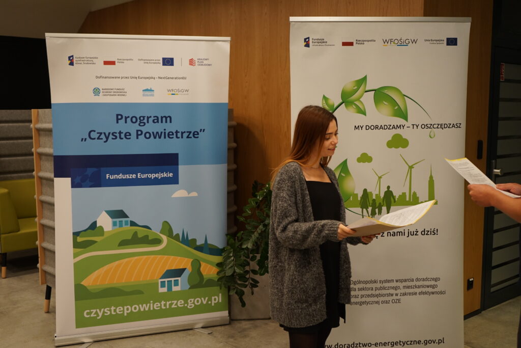 Jedna osoba stoi na tle bannerów reklamowych i omawia wniosek z beneficjentem.