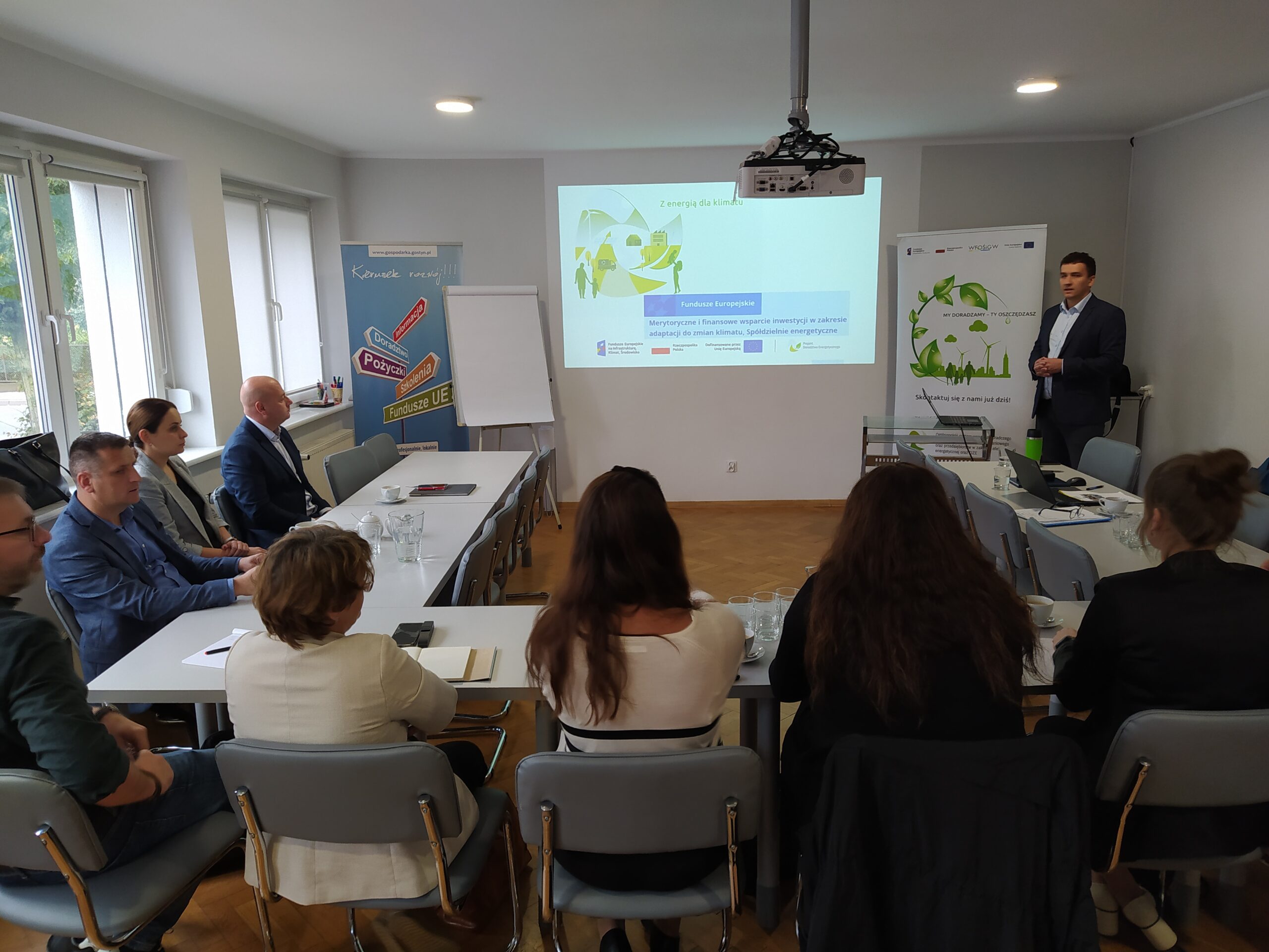 Na zdjęciu wokół stołu siedzą osoby, a na środku na ekranie wyświetlana jest prezentacja, która jest omawiana.