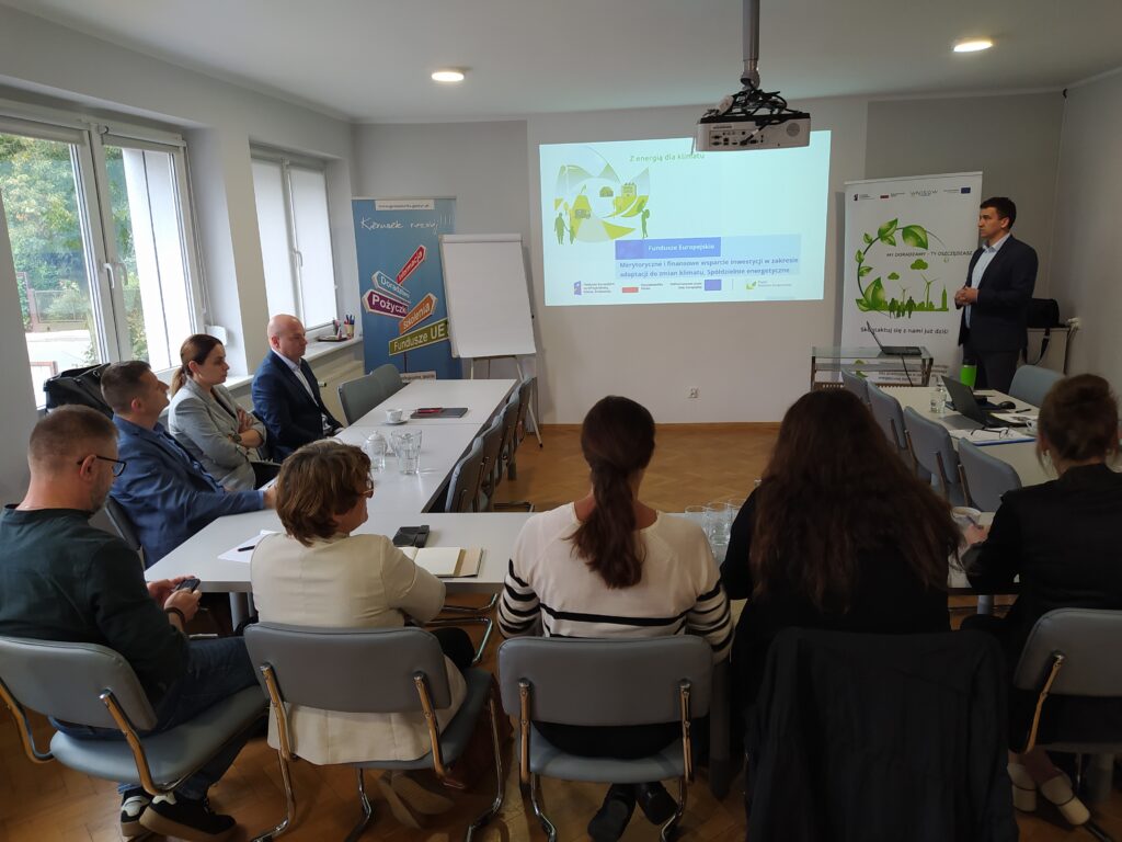 Na zdjęciu wokół stołu siedzą osoby, a na środku na ekranie wyświetlana jest prezentacja, która jest omawiana.