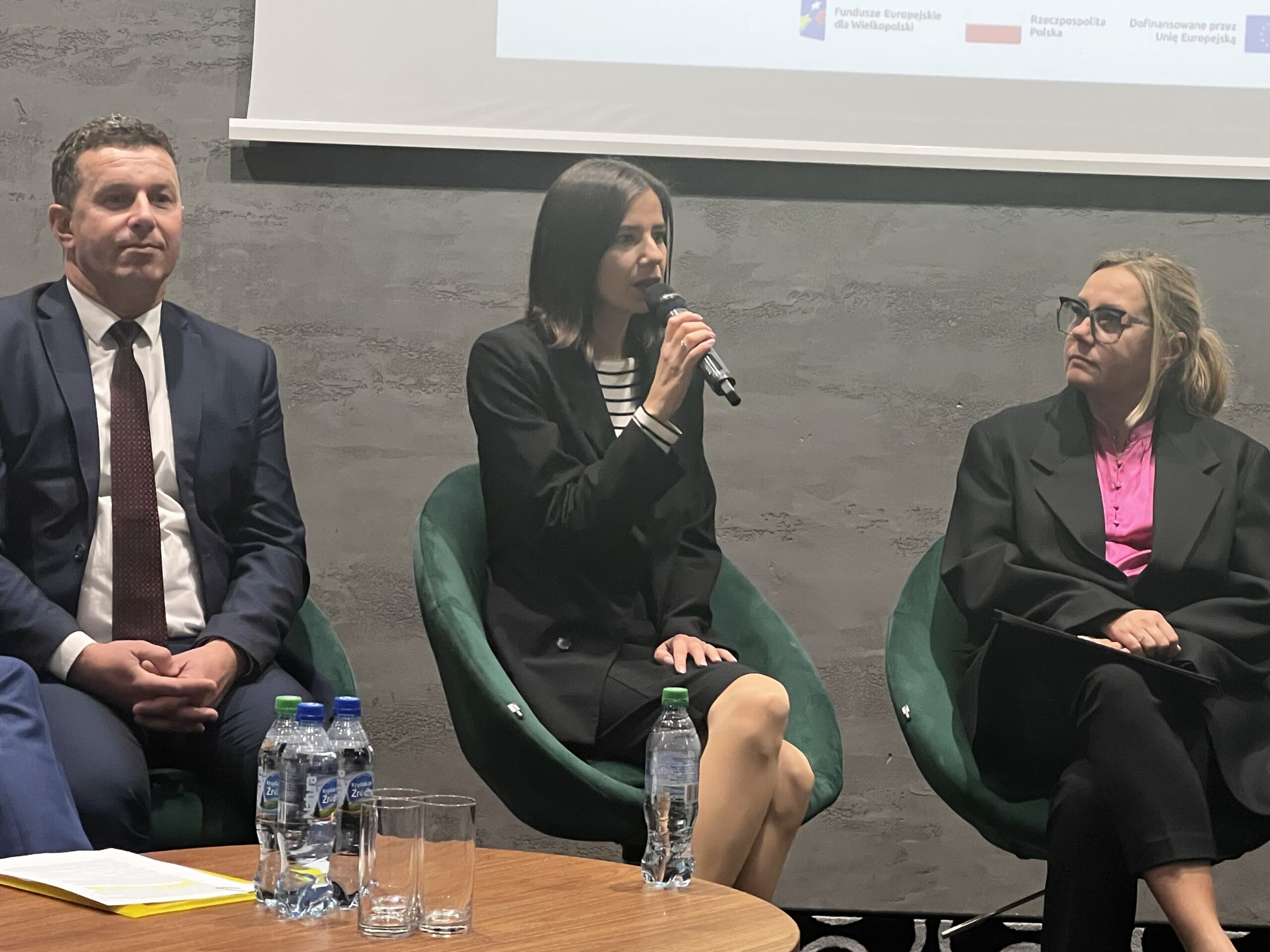 Zdjęcie przedstawia 3 osoby, które siedza na fotelach na tle wyświetlanej na ekranie prezentacji, jedna z osób przemawia do mikrofonu.