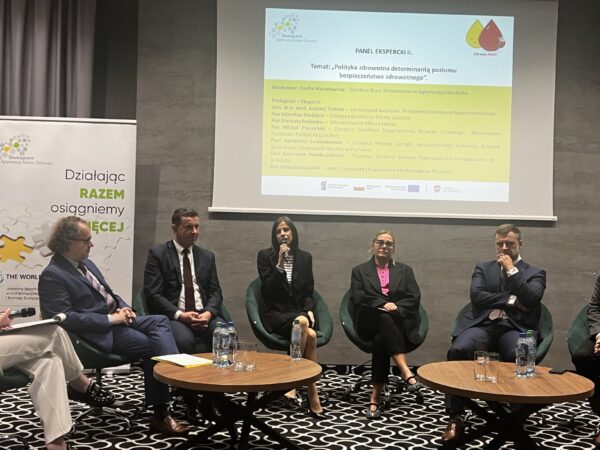 Na sali, na tle wyświetlonej na ekranie prezentacji siedzi 7 osób, które biorą udział w debacie.