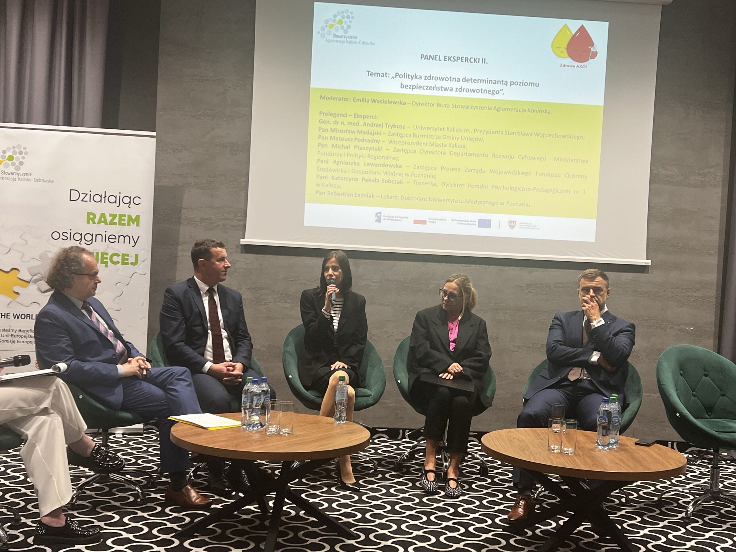 Na sali, na tle wyświetlonej na ekranie prezentacji siedzi 6 osób, które biorą udział w debacie.