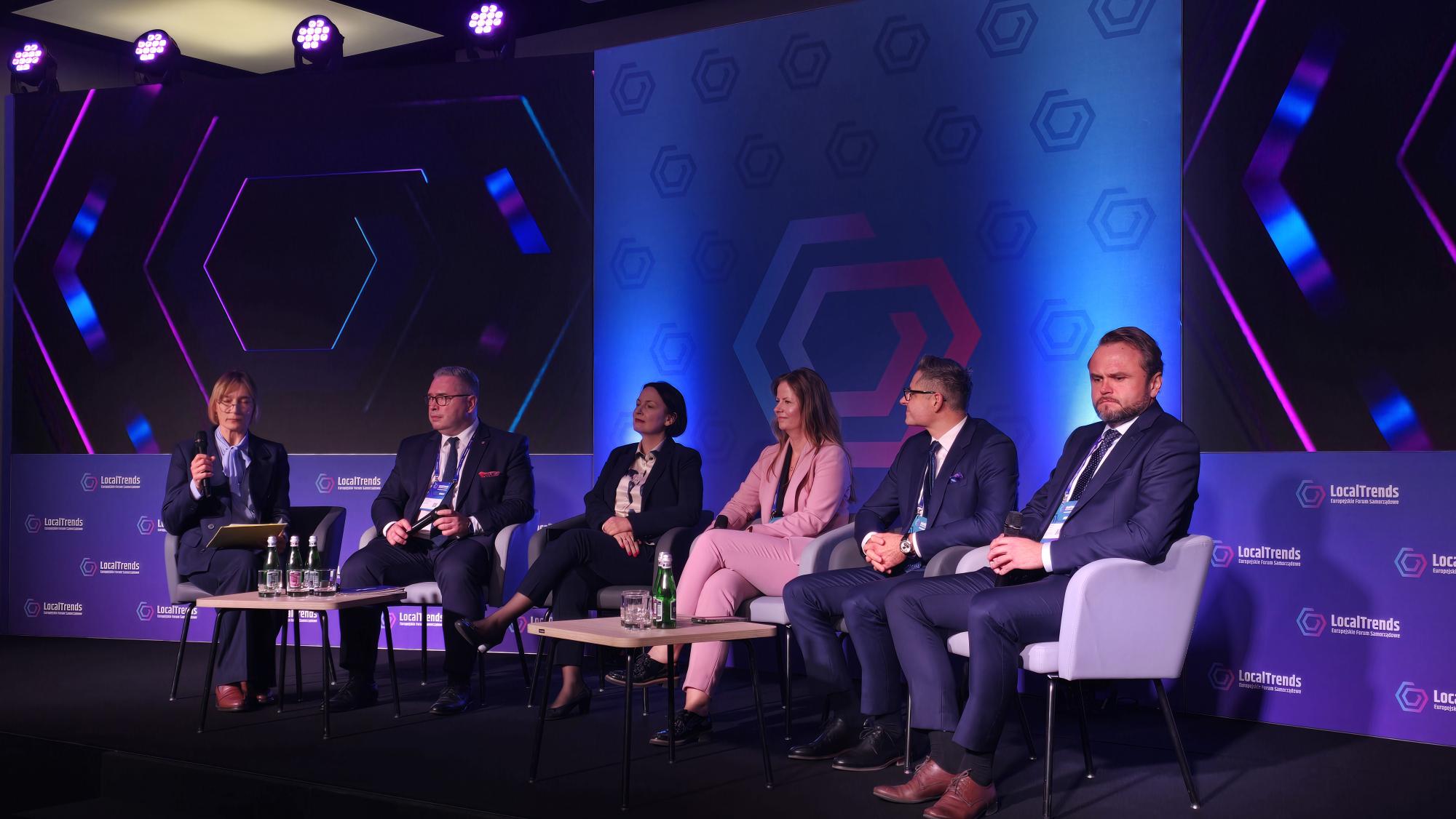 6 osób siedzi na krzesłach na zaaranżowanej podczas wydarzenia scenie, na tle ekranu, na którym wyświetlony jest temat debaty i rozmawia.