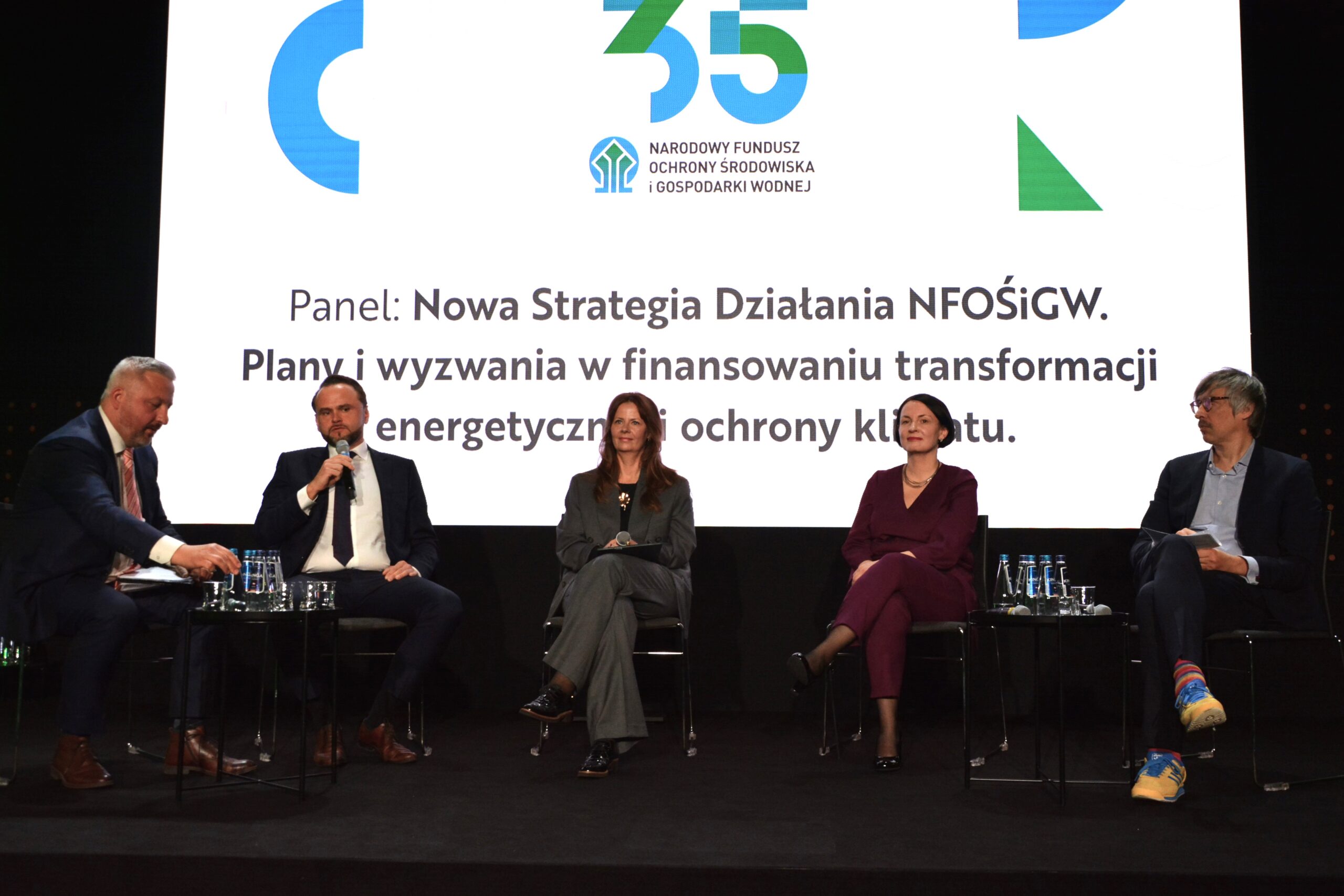 5 osób siedzi na krzesłach na tle telebimu i dyskutują.