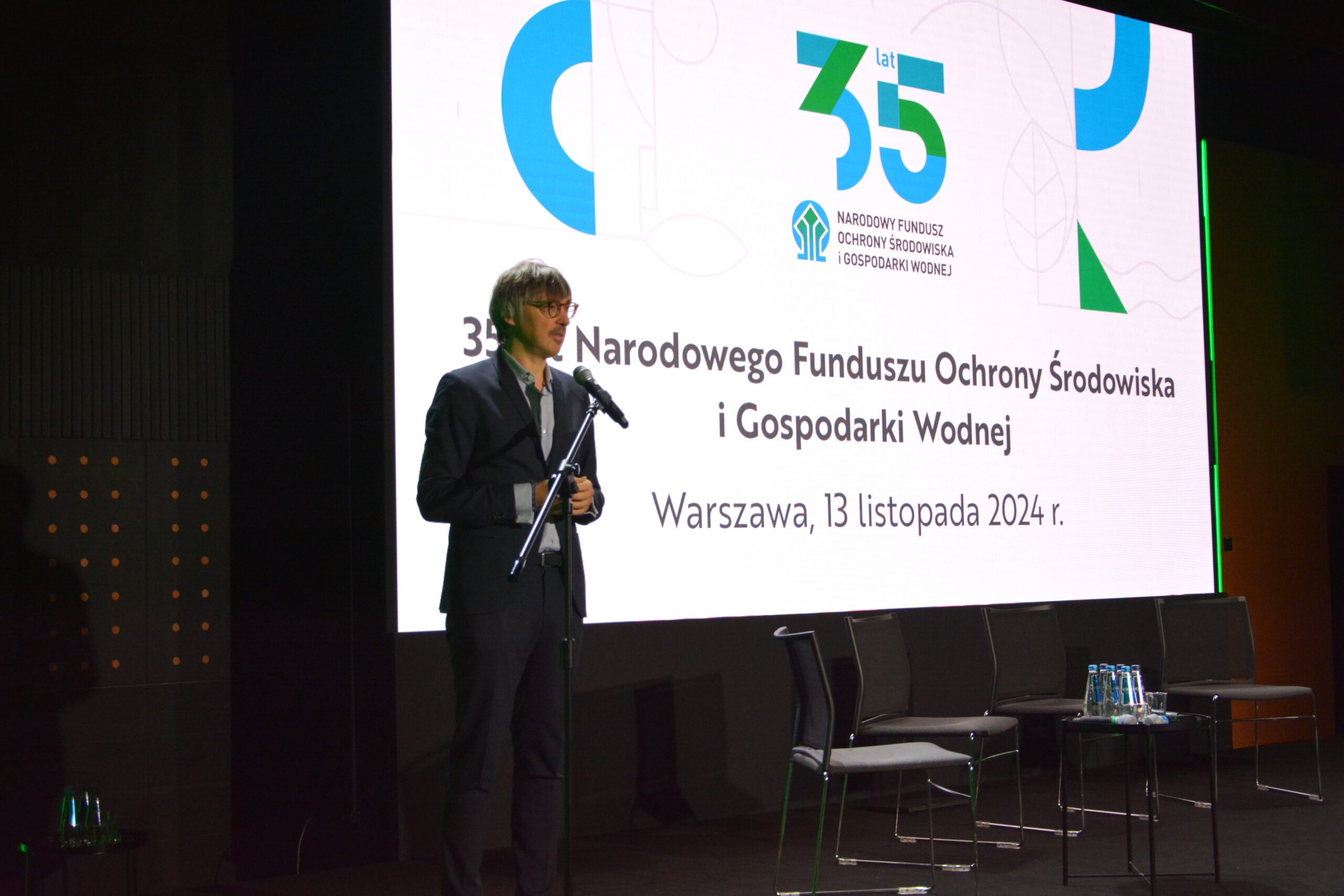 1 osoba stoi na scenie przy mikrofonie na statywie i przemawia.