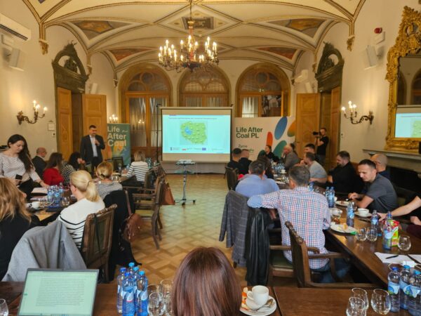 Zdjęcie przedstawia salemw pałacu, w którym odbywa się szkolenie, widać ludzi siedzących na krzesłach patrzących w kierunku wyśwoetlanej prezentacji na ekranie projektora na środku sali.