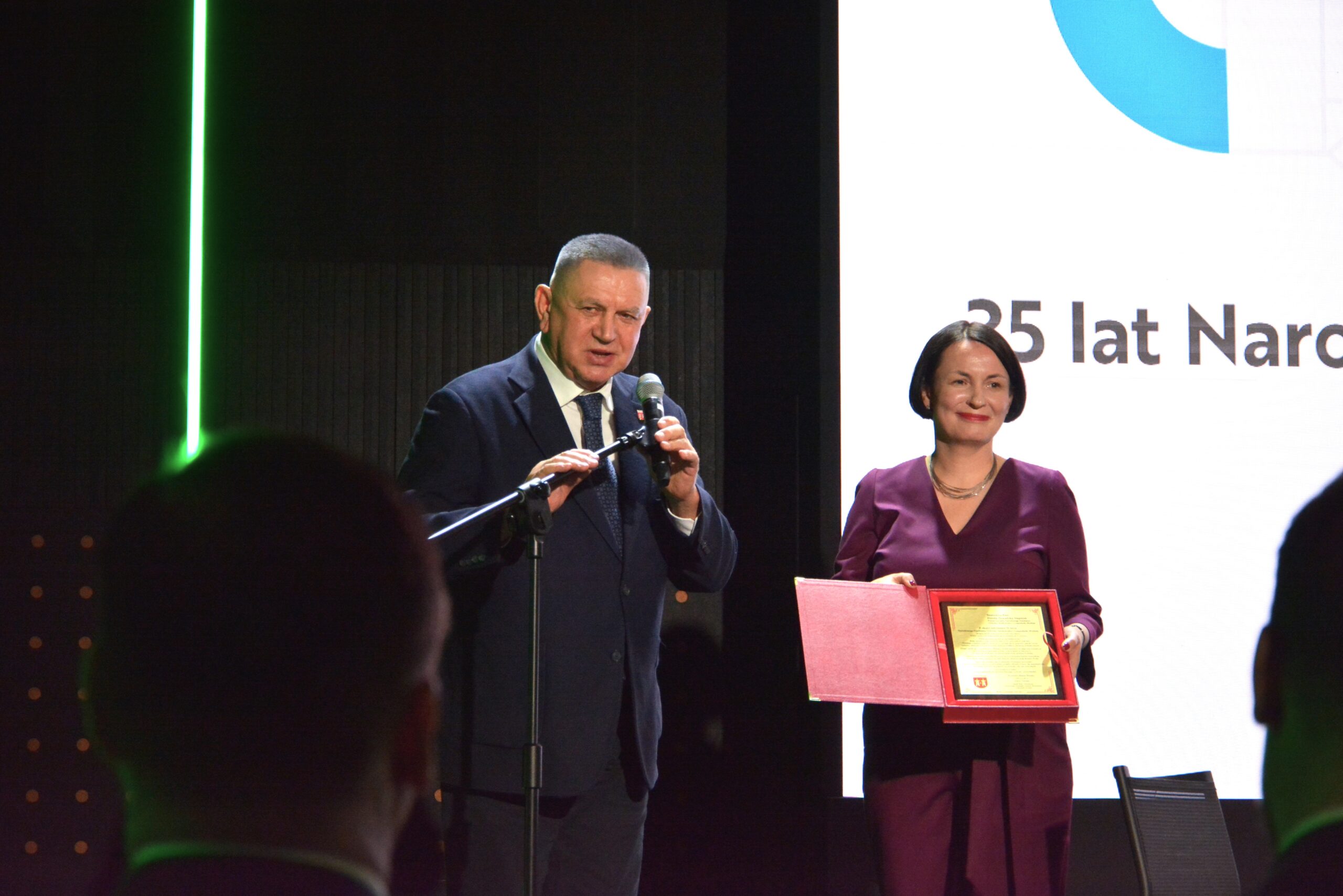 2 osoby stoją na scenie przy mikrofonie umieszczonym na statywie i jedna z nich przemawia, a druga prezentuje otrzymaną nagrodę.