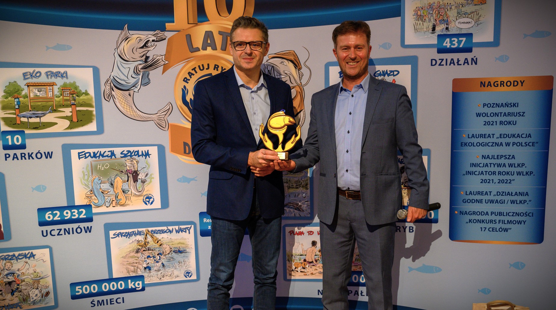 Na tle ścianki reklamowej stoi Prezes Zarządu WFOŚiGW w Poznaniu, który w ręku trzyma otrzymaną oraz Prezes Fundacji "Ratuj Ryby".