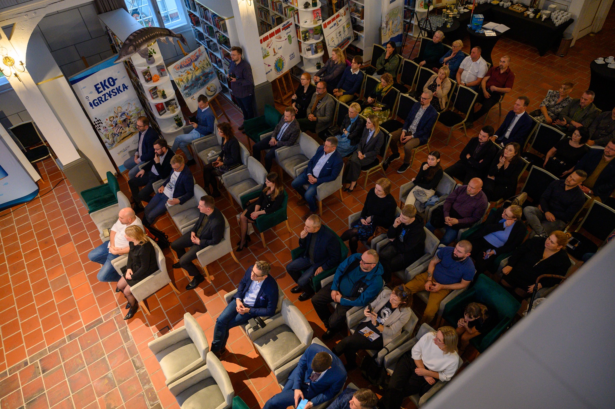 Zdjęcie zrobione jest z góry i przedstawia widok na salę, na której na krzesłach siedzą zaproszeni goście.