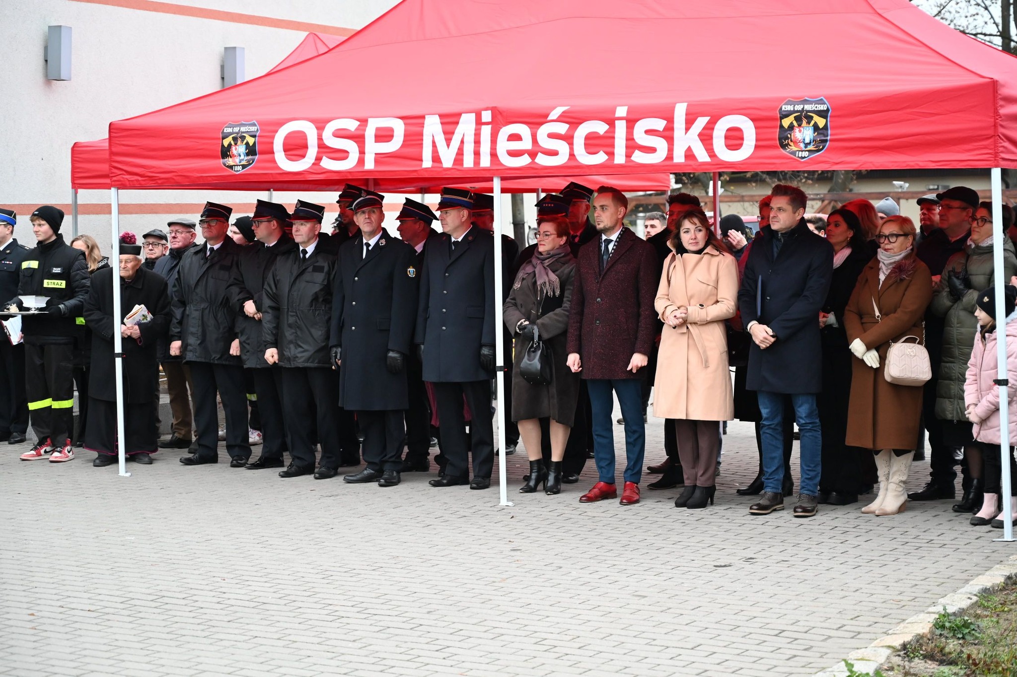 Na zdjęciu, na placu, pod zadaszeniem z namiotu OSP Mieścisko, stoi grupa ludzi - zaproszeni goście na powitanie nowego wozu strażackiego w gminie Mieścisko.