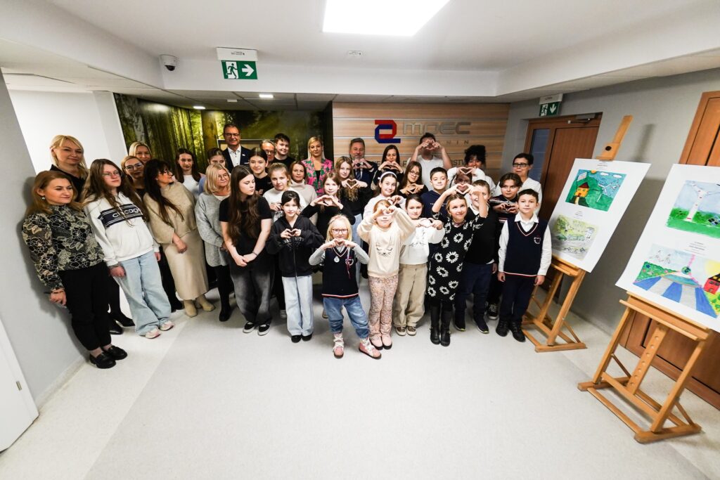 W holu siedziby firmy MPEC Konin stoi duża grupa osób i pozuje do zdjęcia pośród wystawy złożonej z prac zgłoszonych do konkursu.