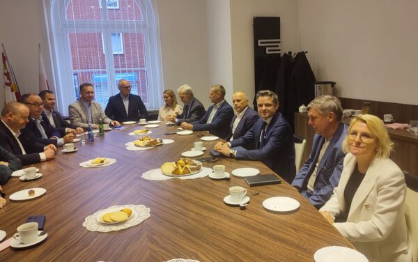 Zdjęcie przedstawia 13 osób, które siedzą wokół owalnego stołu i prowadzą rozmowę.