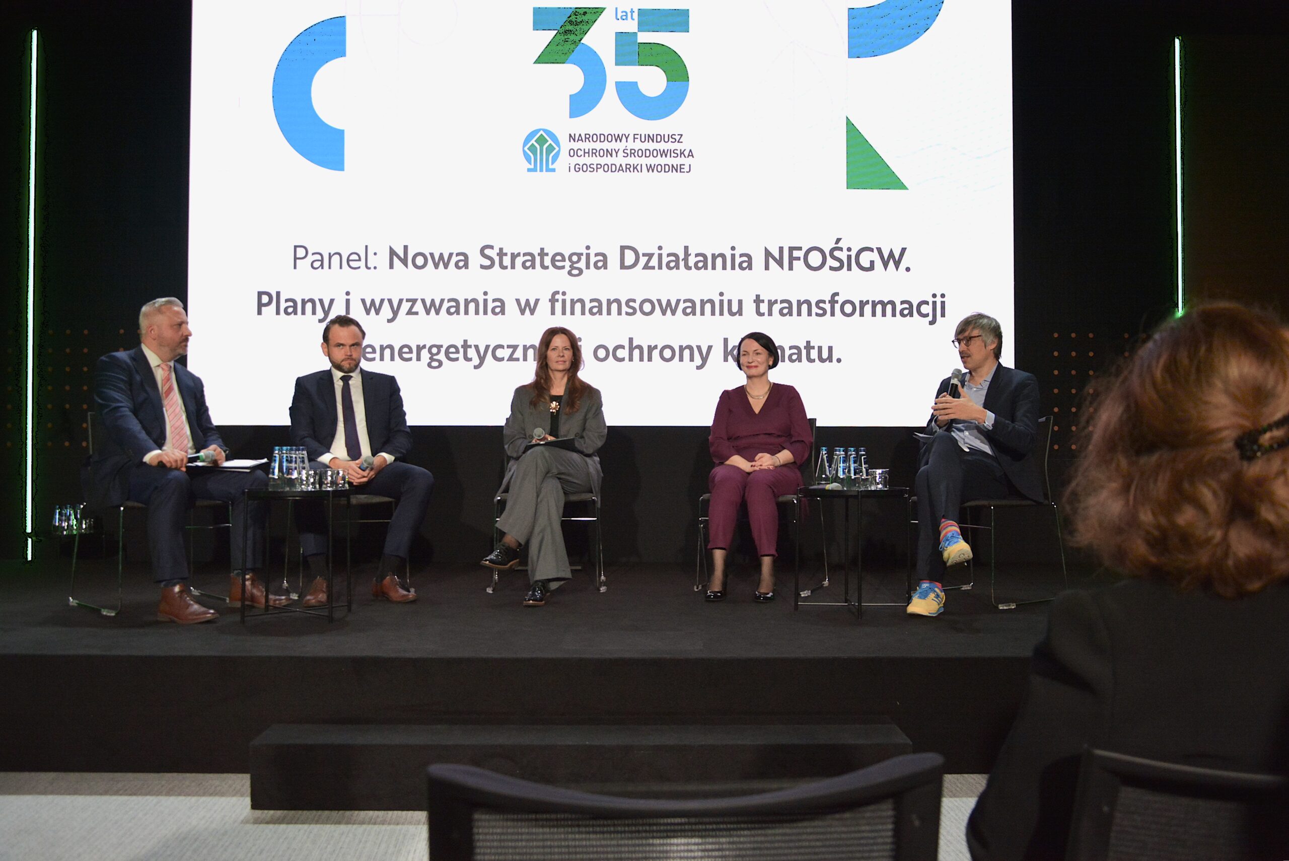 5 osób siedzi na krzesłach na scenie na tle telebimu i dyskutują.