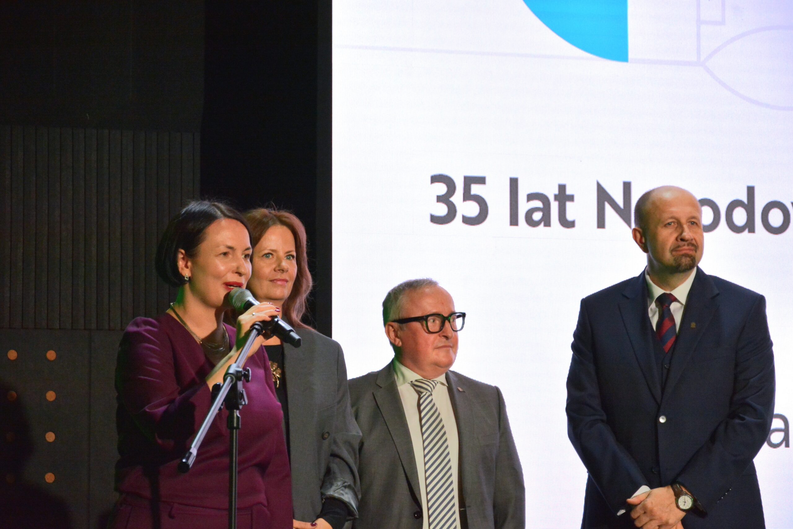 4 osoby stojąna scenie, jedna z nich przemawia do mikrofonu umieszczonego na statywie, a pozostałe 3 osoby stoją obok i przysłuchują się.