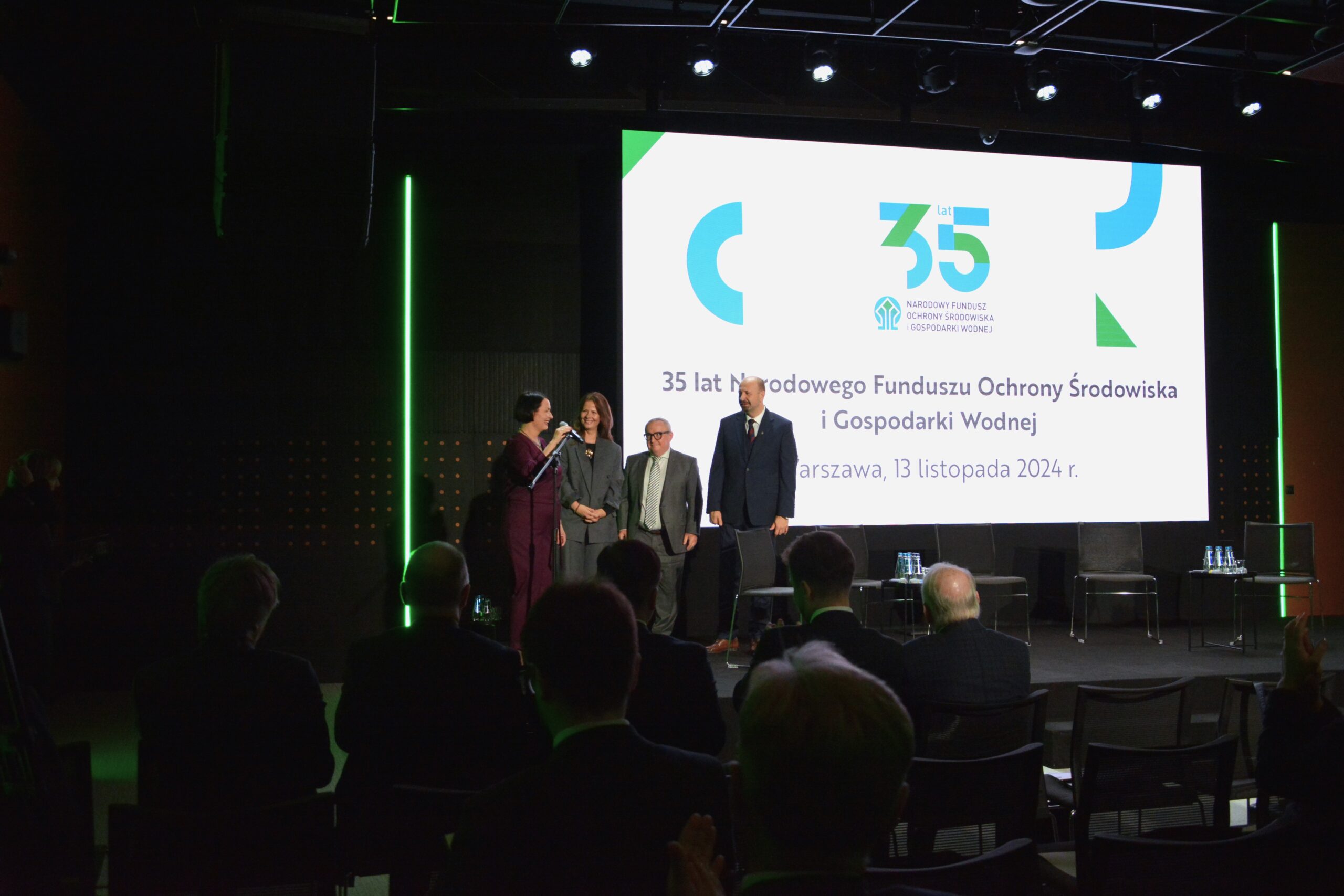 4 osoby stoją na scenie, jedna z nich przemawia do mikrofonu umieszczonego na statywie, a pozostałe 3 osoby stoją obok i przysłuchują się oraz uśmiechają.