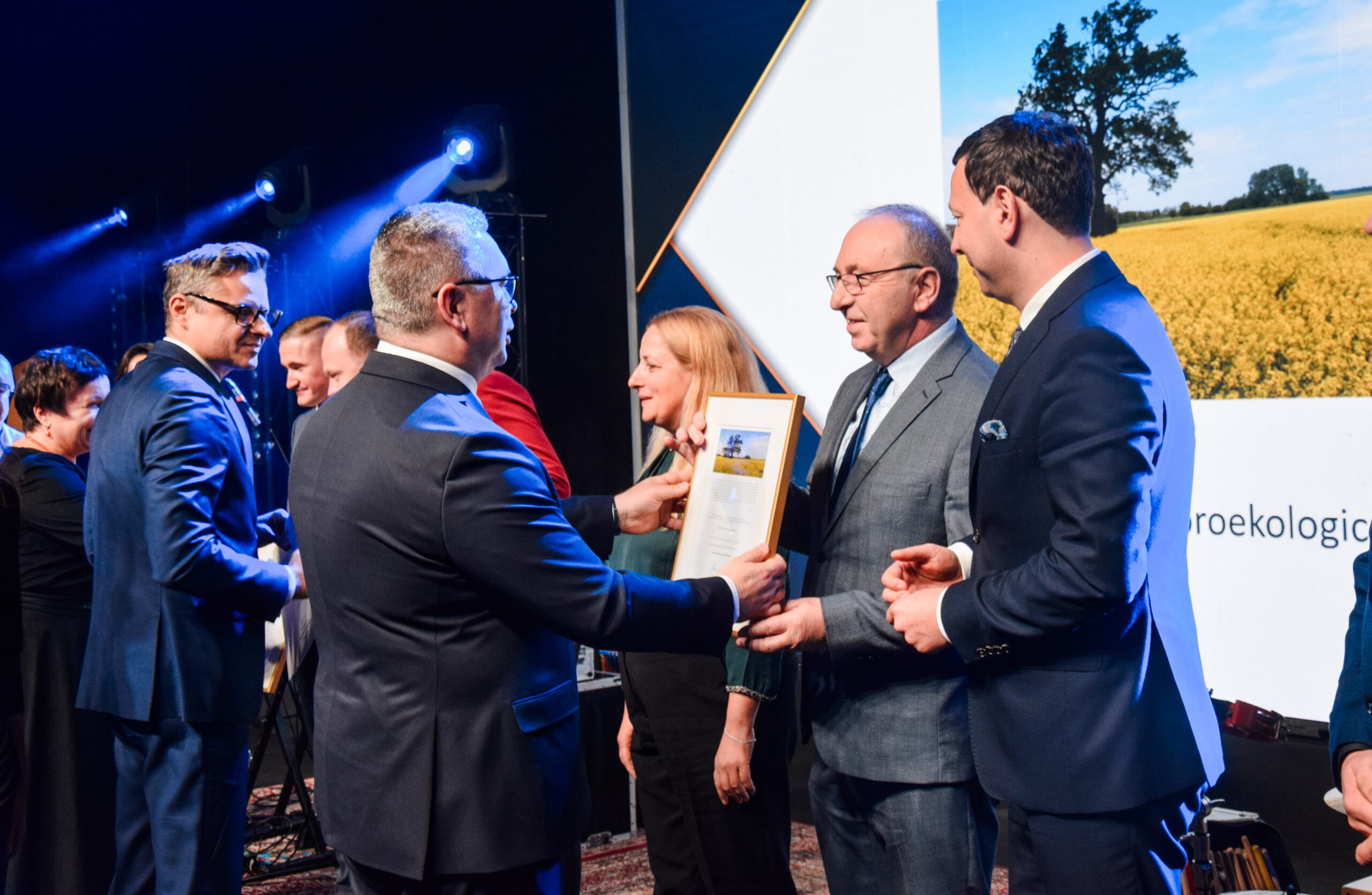 Na scenie stoi kilka osób i kolejne osoby wręczają nagrody oraz dyplomy.