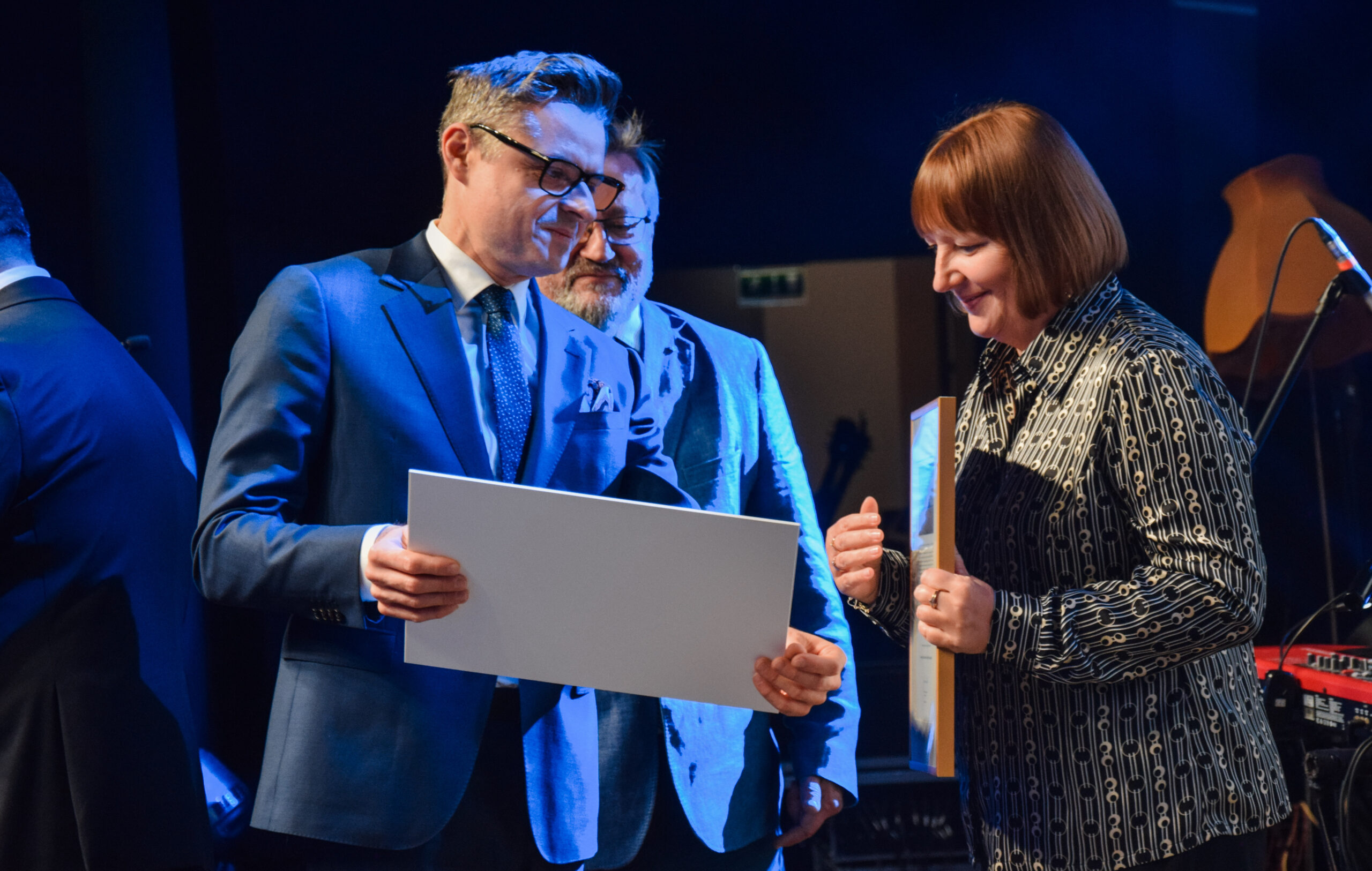 Na scenie stoi kilka osób i kolejne osoby wręczają nagrody oraz dyplomy.