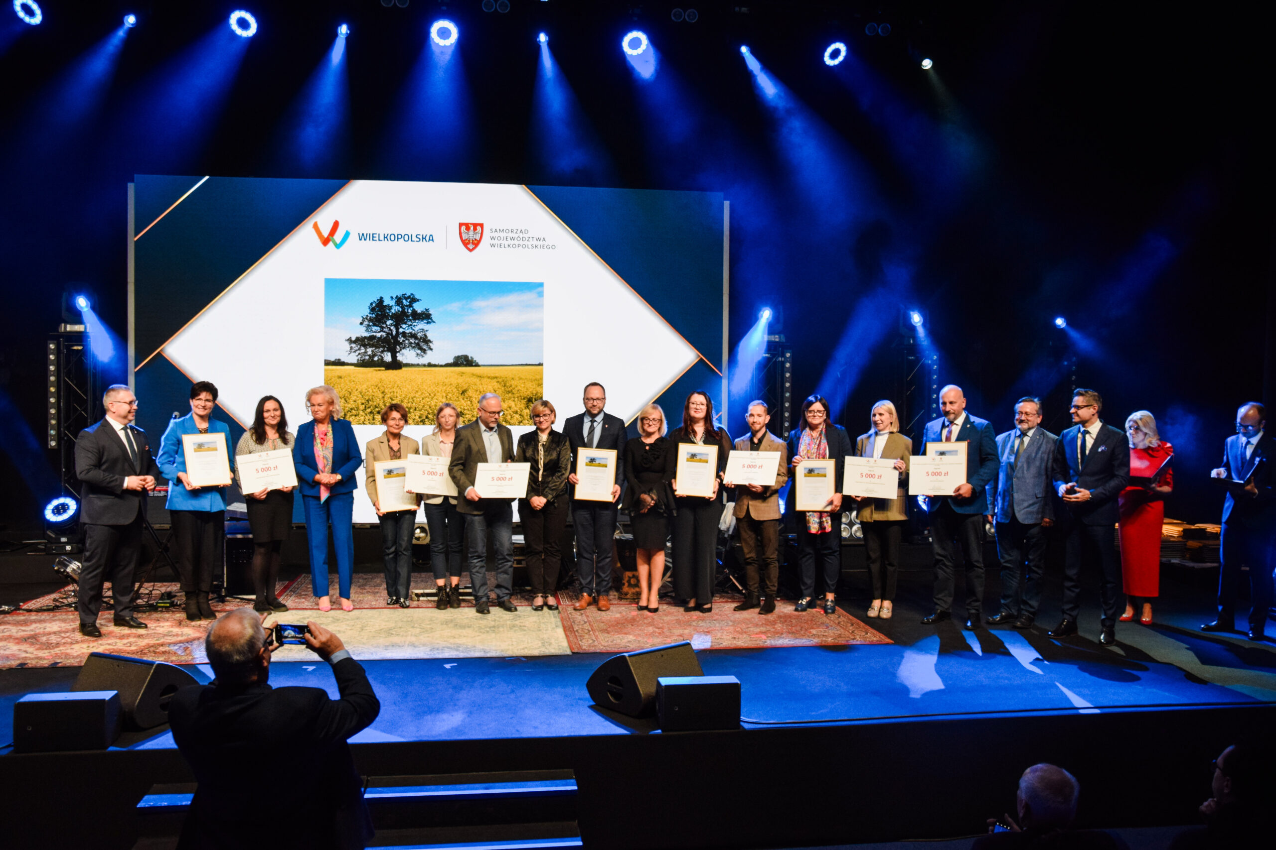 Na zdjęciu stoi duża grupa nagrodzonych osób i wspólnie pozuje do zdjęcia wraz z nagrodami.
