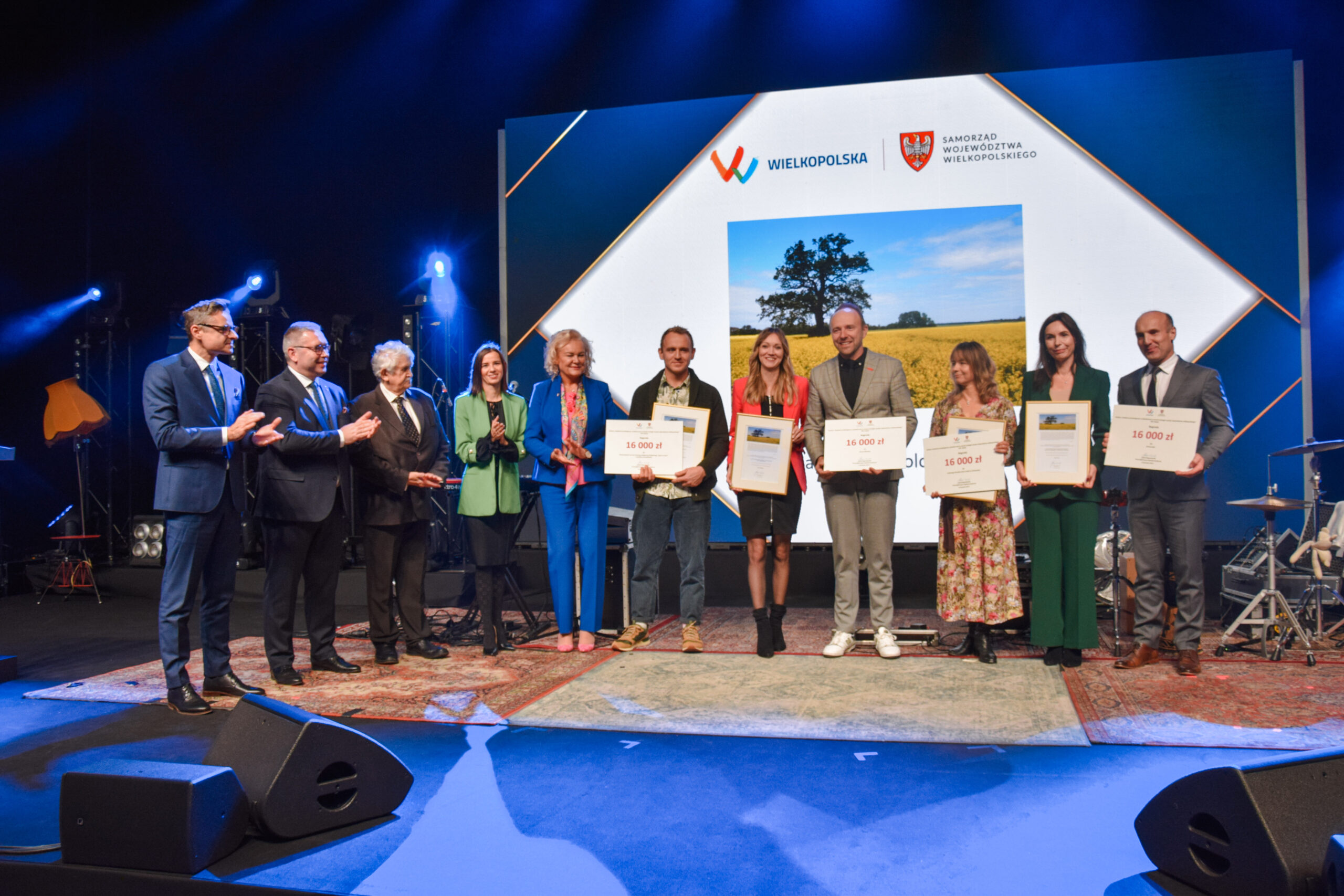 Na zdjęciu stoi duża grupa nagrodzonych osób i wspólnie pozuje do zdjęcia wraz z nagrodami.