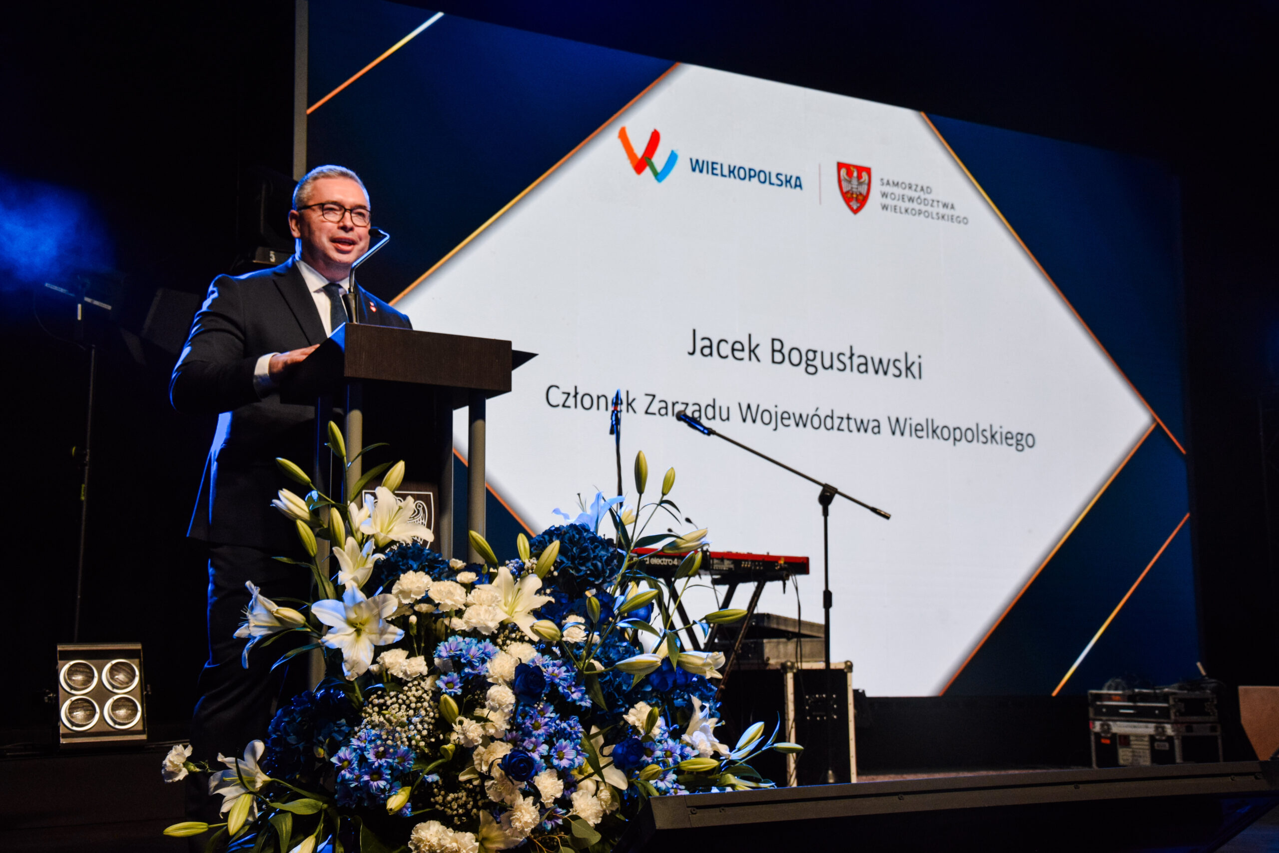 Na scenie przy mównicy stoi jedna osoba i wypowiada się.