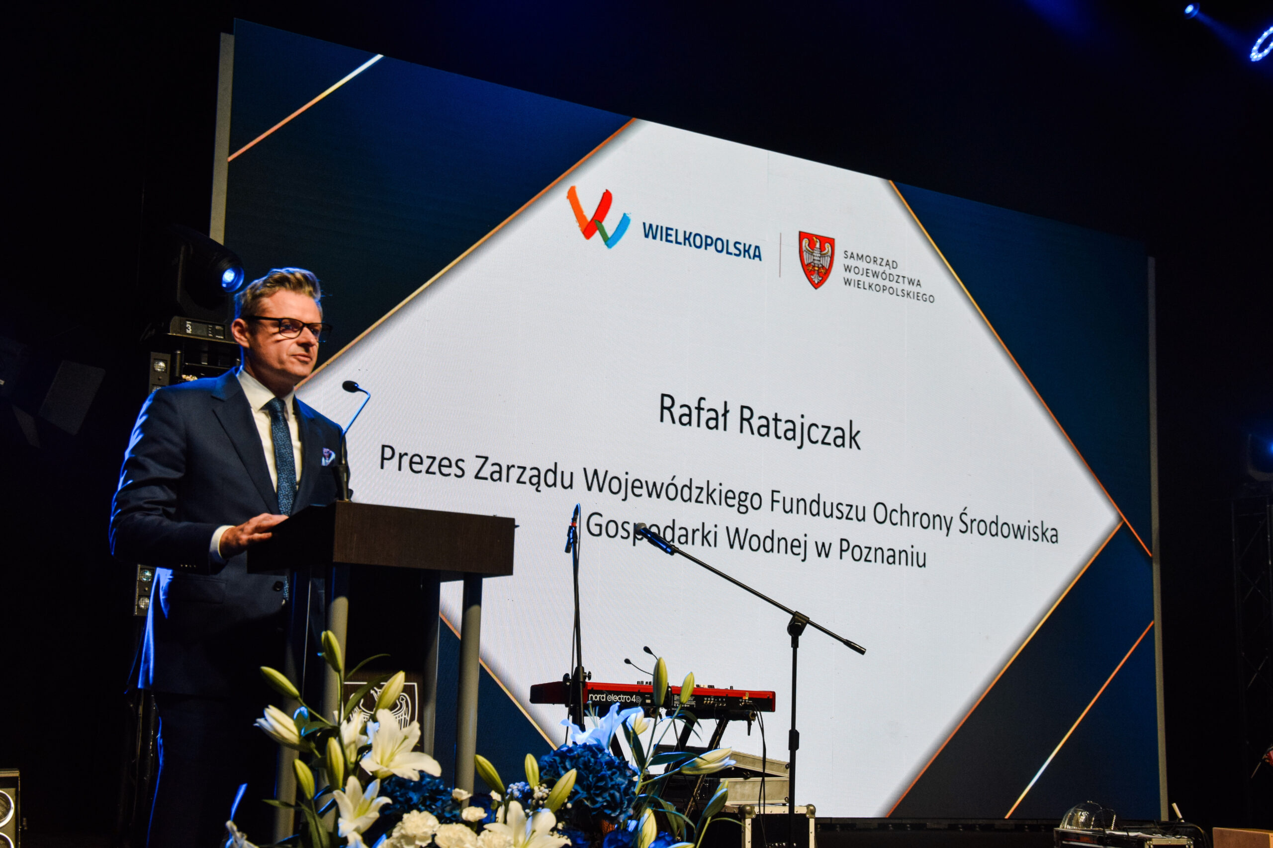 Na scenie przy mównicy stoi jedna osoba i wypowiada się.