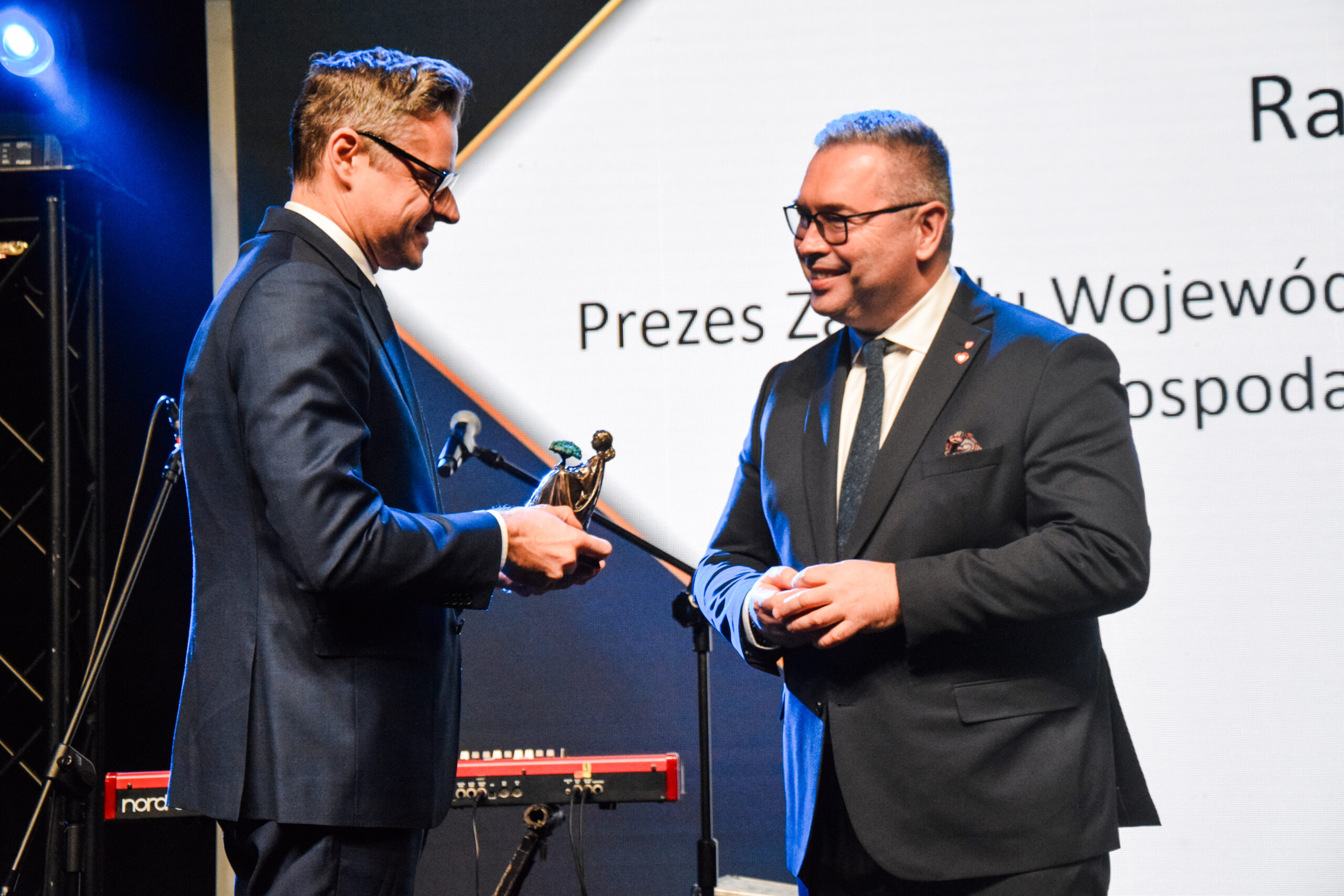 Na zdjęciu na scenie stoją 2 osoby, jedna z nich wręcza drugiej statuetkę.