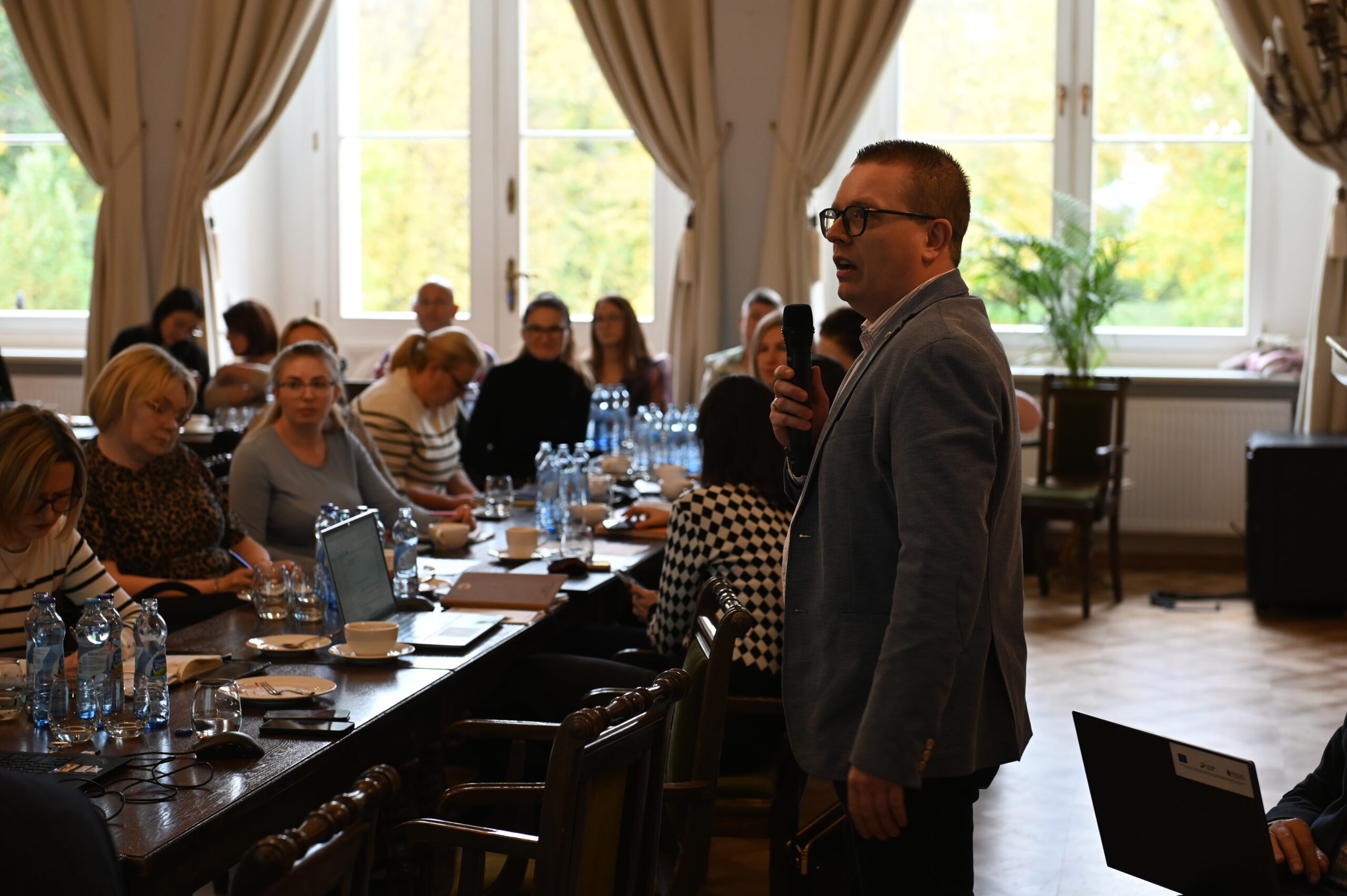 Zdjęcie przedstawia osobę prowadzącą szkolenia, która stoi przed stołami i mówi do mikrofonu.