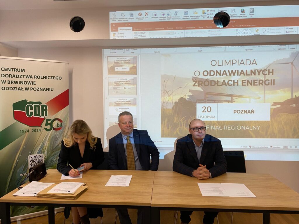 Zdjęcie przedstawia 3osoby siedzące za stołem na tle wyświetlonego ekranu z projektora.