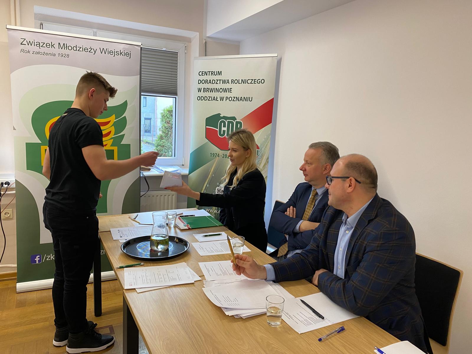 Na zdjęciu widać trzy osoby siedzące za stołem, a przed nimi stoi jedna osoba.