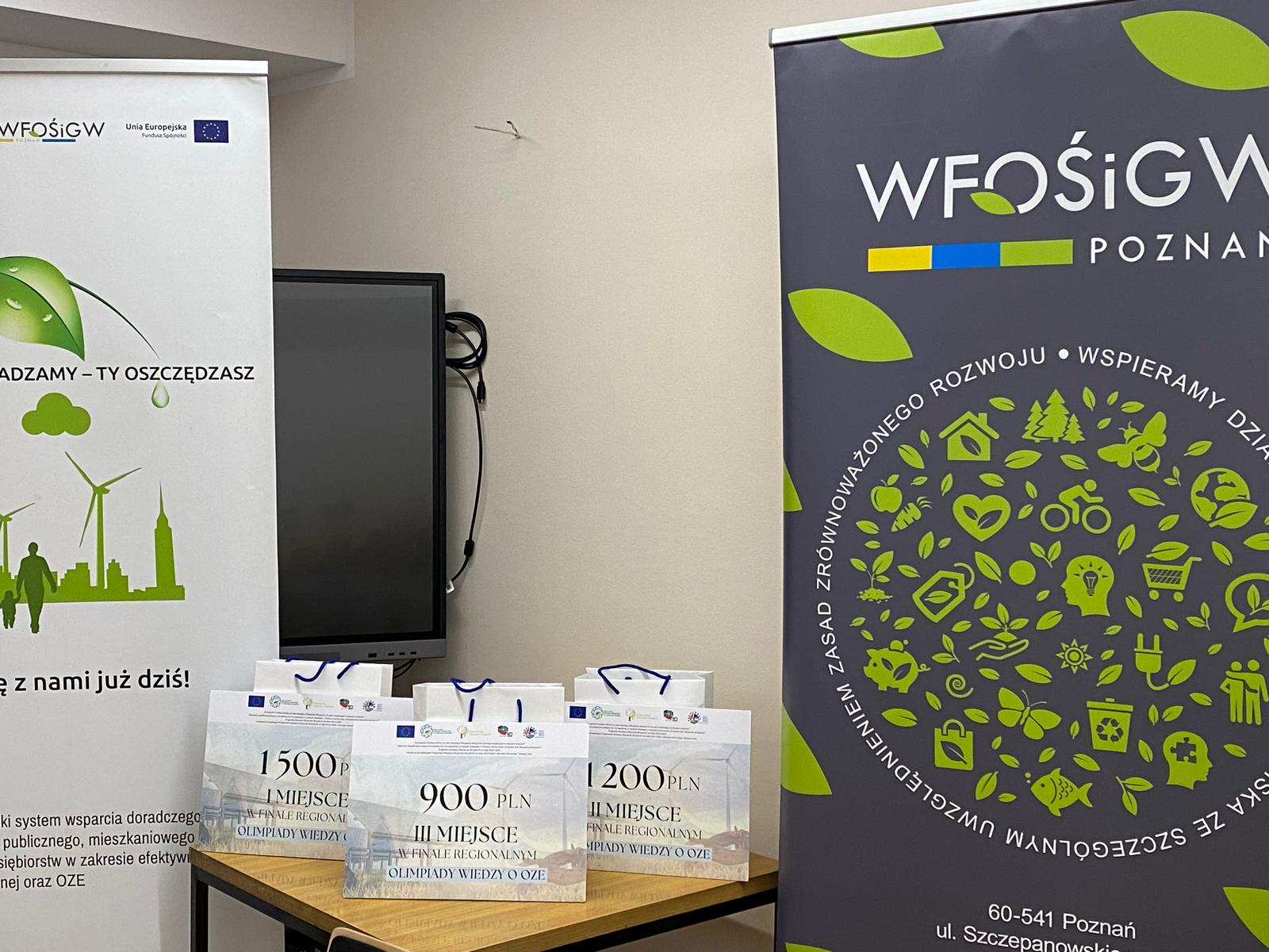 Zdjęcie przedstawia widok na stole, na którym stoją torby z upominkami dla uczestników oraz po obu stronach stoją rollupy.