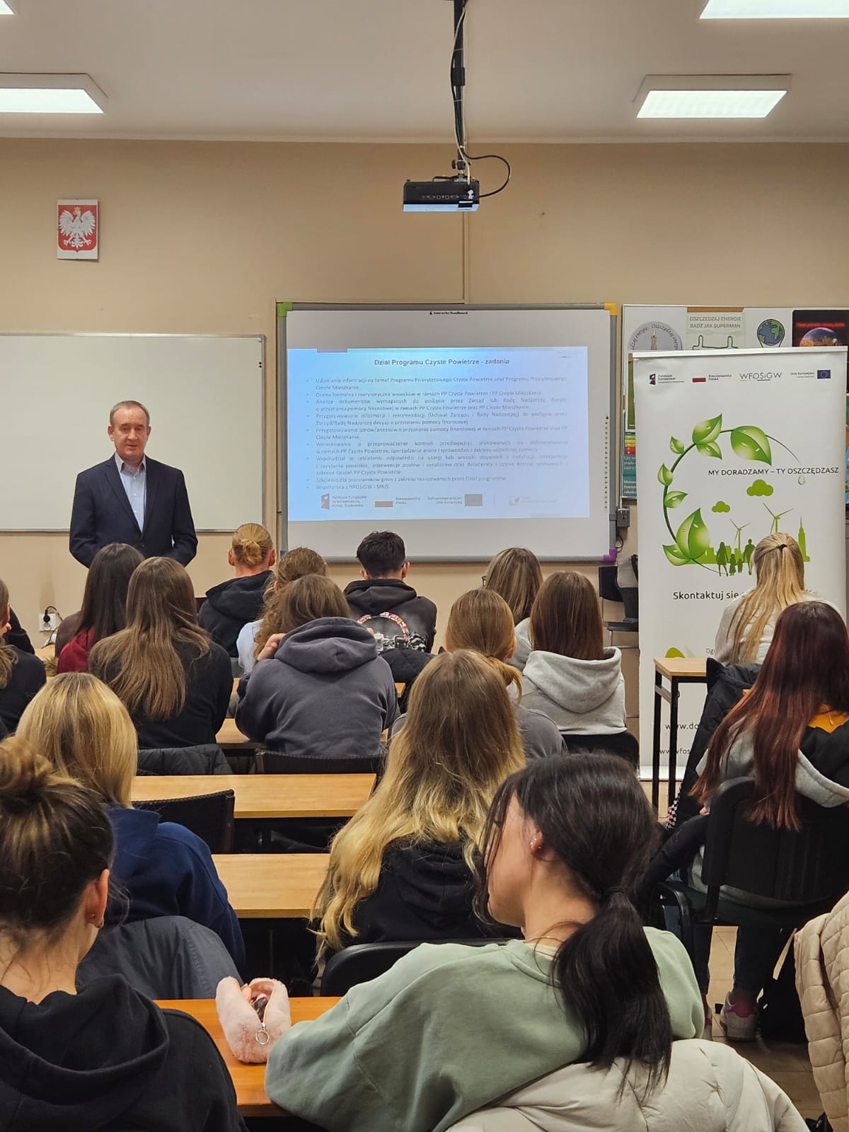 Zdjęcie przedstawia salę lekcyjną, na krzesłach w ławkach siedzą uczniowie, a na przodzie sali obok biurka stoi osoba i omawia wyświetloną na ekranie projektora prezentację.