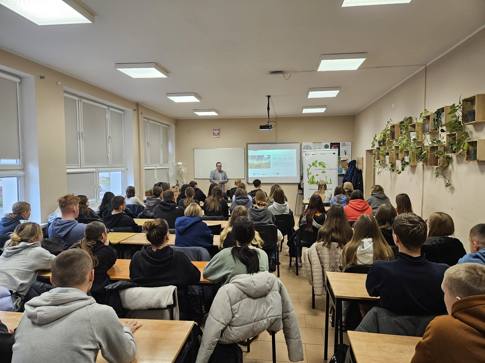 Zdjęcie przedstawia salę lekcyjną, na krzesłach w ławkach siedzą uczniowie, a za biurkiem stoi osoba i omawia wyświetloną na ekranie projektora prezentację.