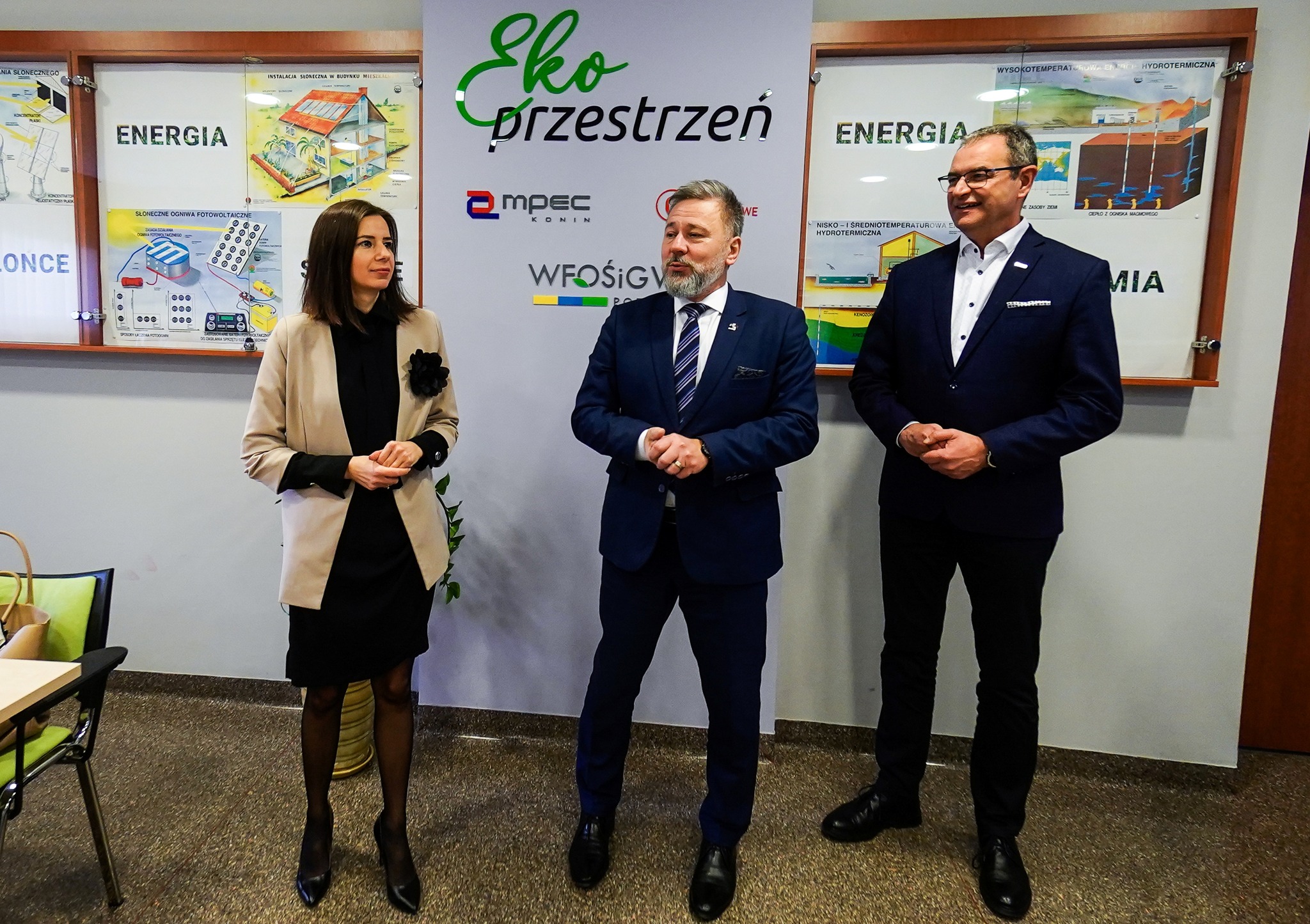 Na tle tablic z ogłoszeniami stoją 3 osoby.