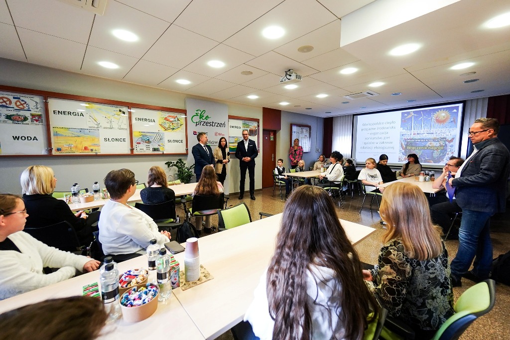 Na sali, za długimi stołami siedzą uczniowie biorący udział w konkursie, a na środku stoją 3 osoby.