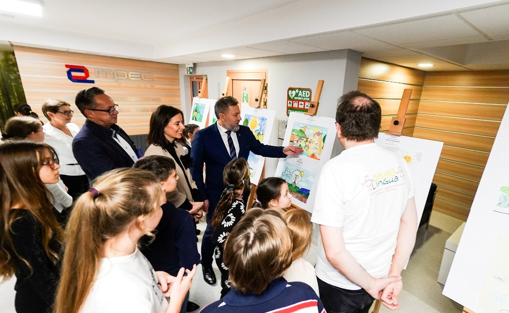 W holu siedziby firmy stoi duża grupa osób i podziwia prace plastyczne zamieszczone na sztalugach.