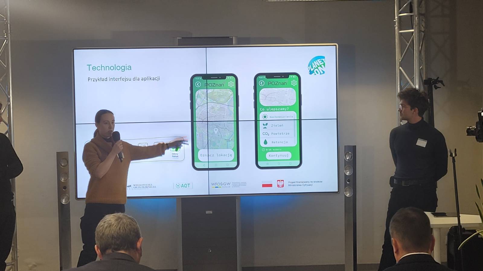 Przed ekranem z wyświetloną prezentacją stoją 2 osoby, jedna z nich omawia prezentację.