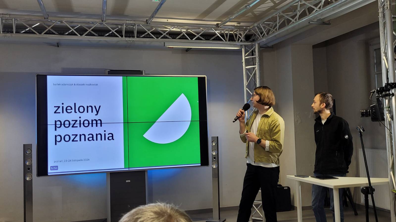 Przed ekranem z wyświetloną prezentacją stoi jedna osoba i omawia prezentację, korzystając z mikrofonu.