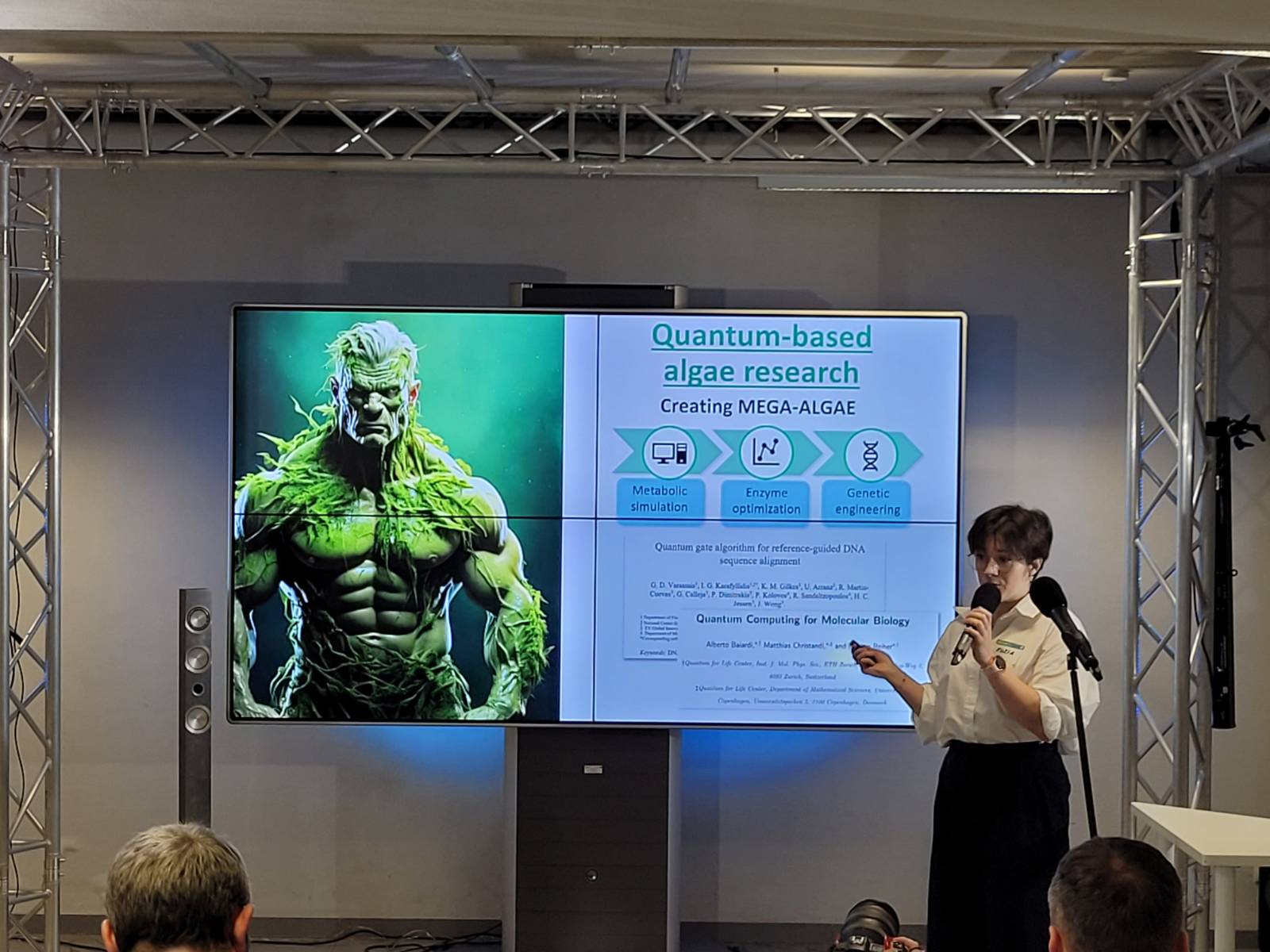 Przed ekranem z wyświetloną prezentacją stoi jedna osoba i omawia prezentację, korzystając z mikrofonu.