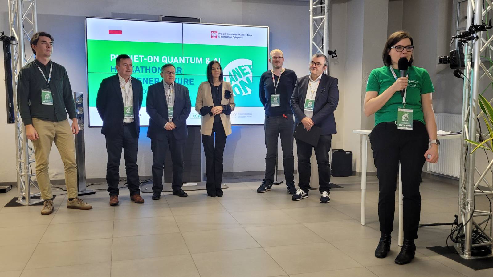 Na zdjęciu na tle ekranu, na którym wyświetlane były prezentacje, stoją osoby będące w jury, a osoba prowadząca wydarzenie przemawia przez mikrofon.