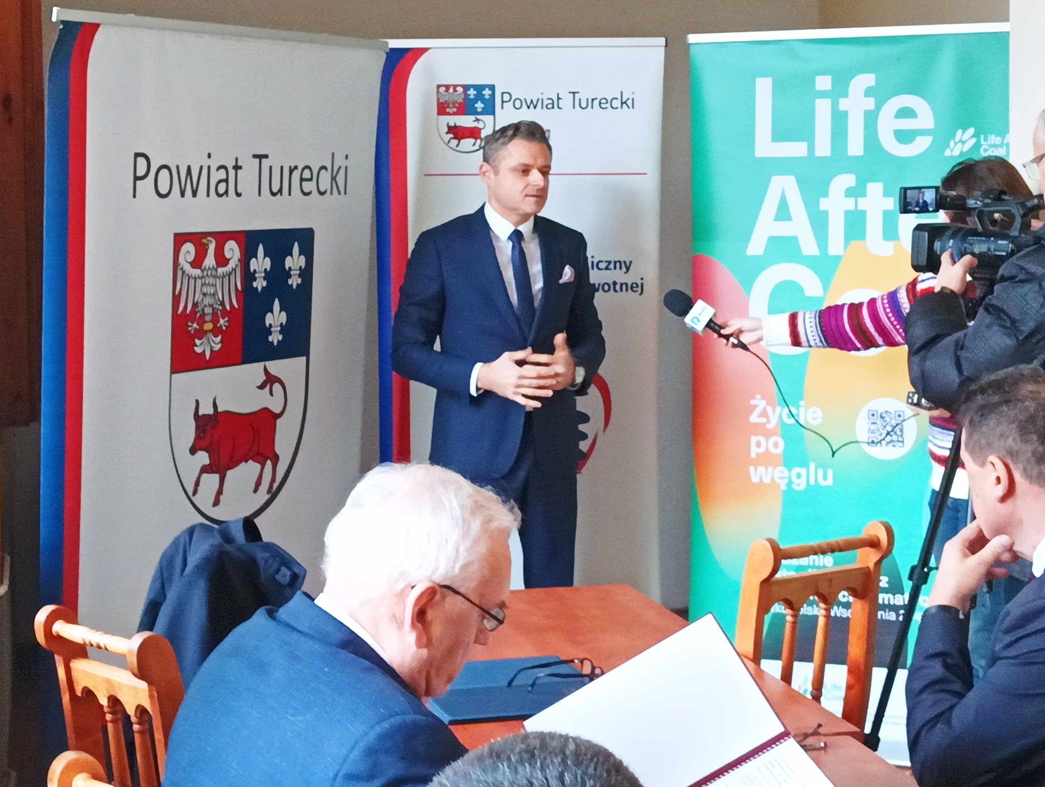 Jedna osoba stoi i mówi do kamery na tle bannerów reklamowych.